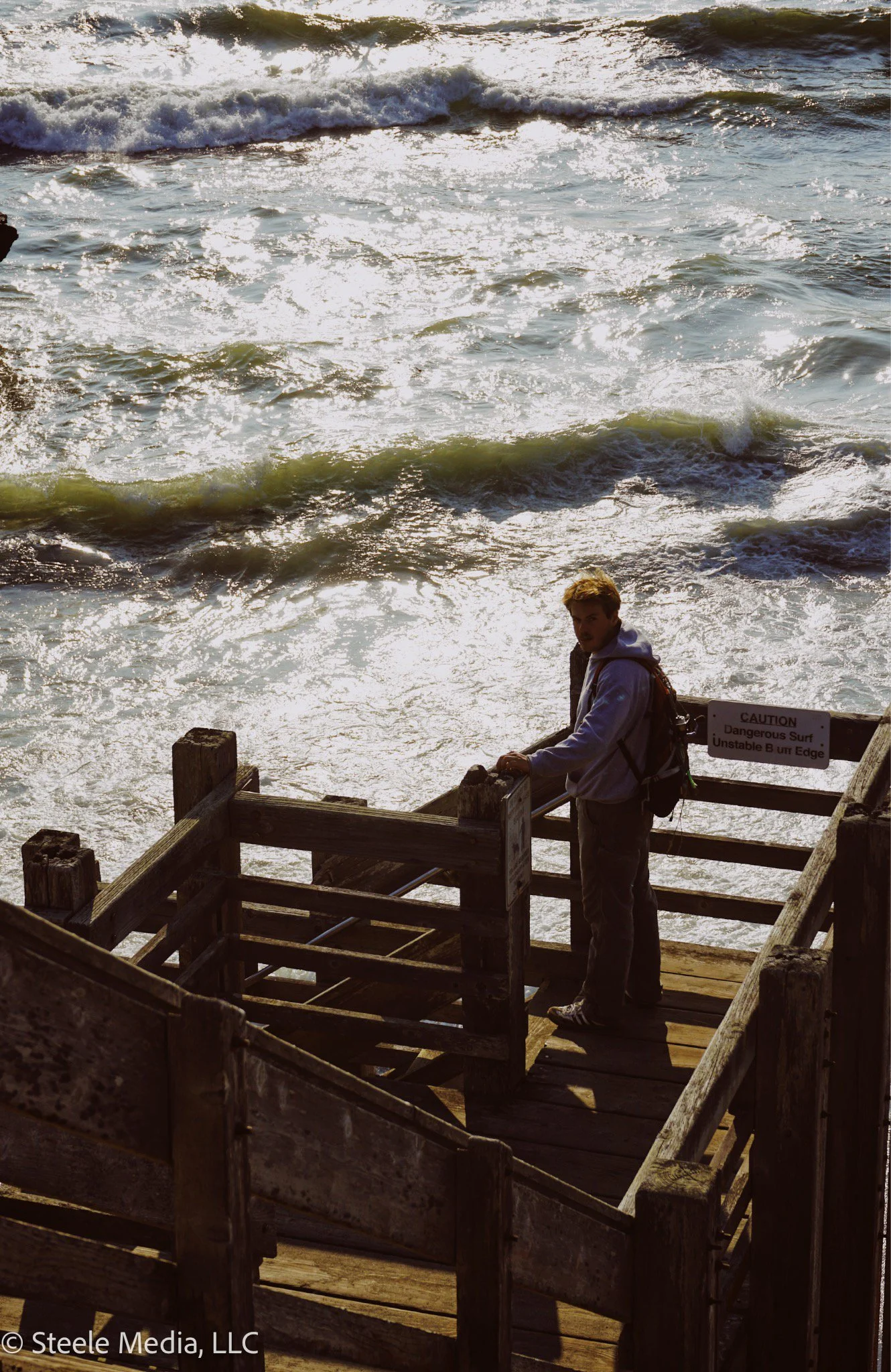 A person standing on a wooden pier looking at the ocean waves. The man is wearing a light-colored hoodie, pants, and has a backpack. There is a sign warning about dangerous surf and unstable edge.