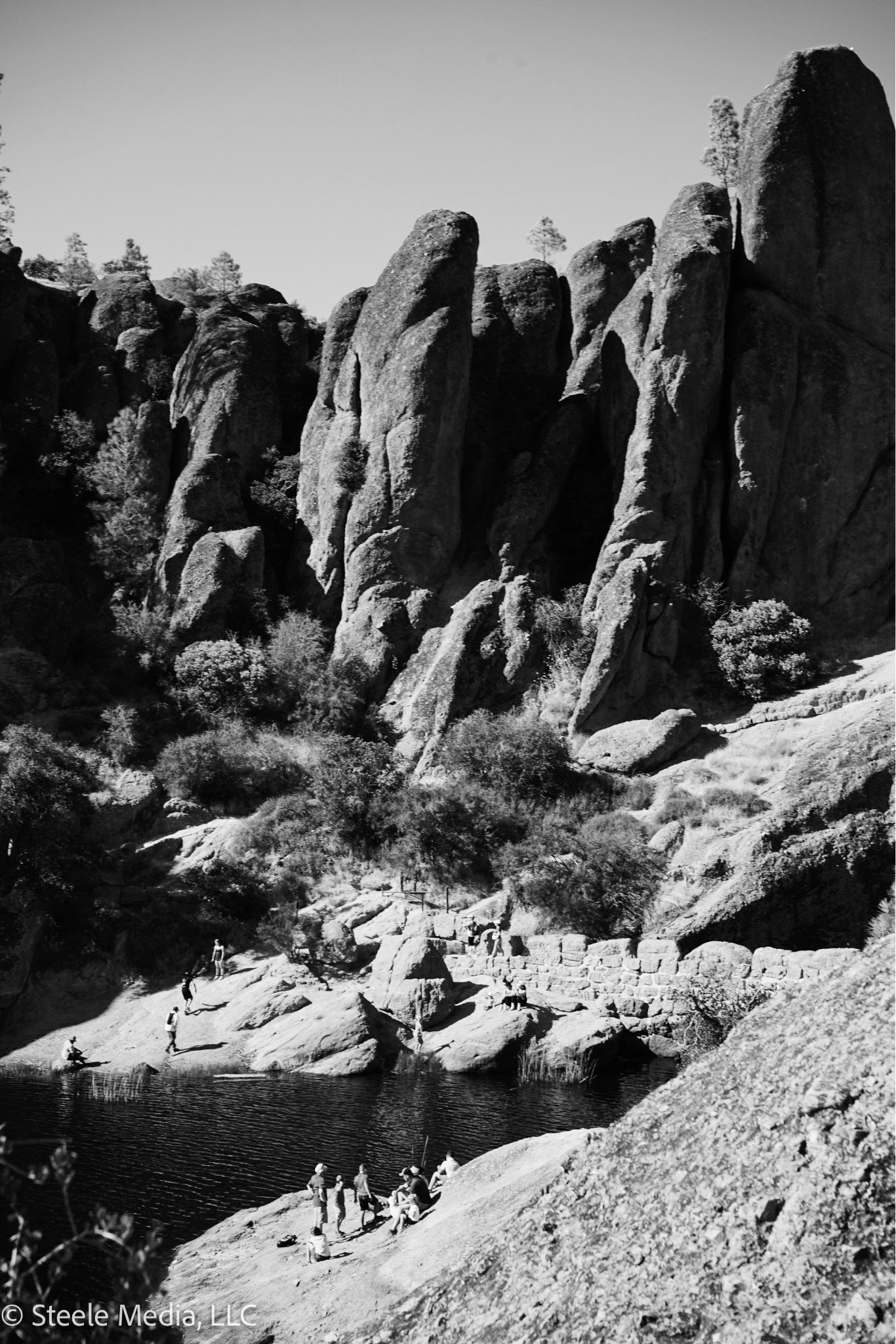People sitting and standing near a body of water surrounded by large rock formations and trees, with rugged terrain in the background.