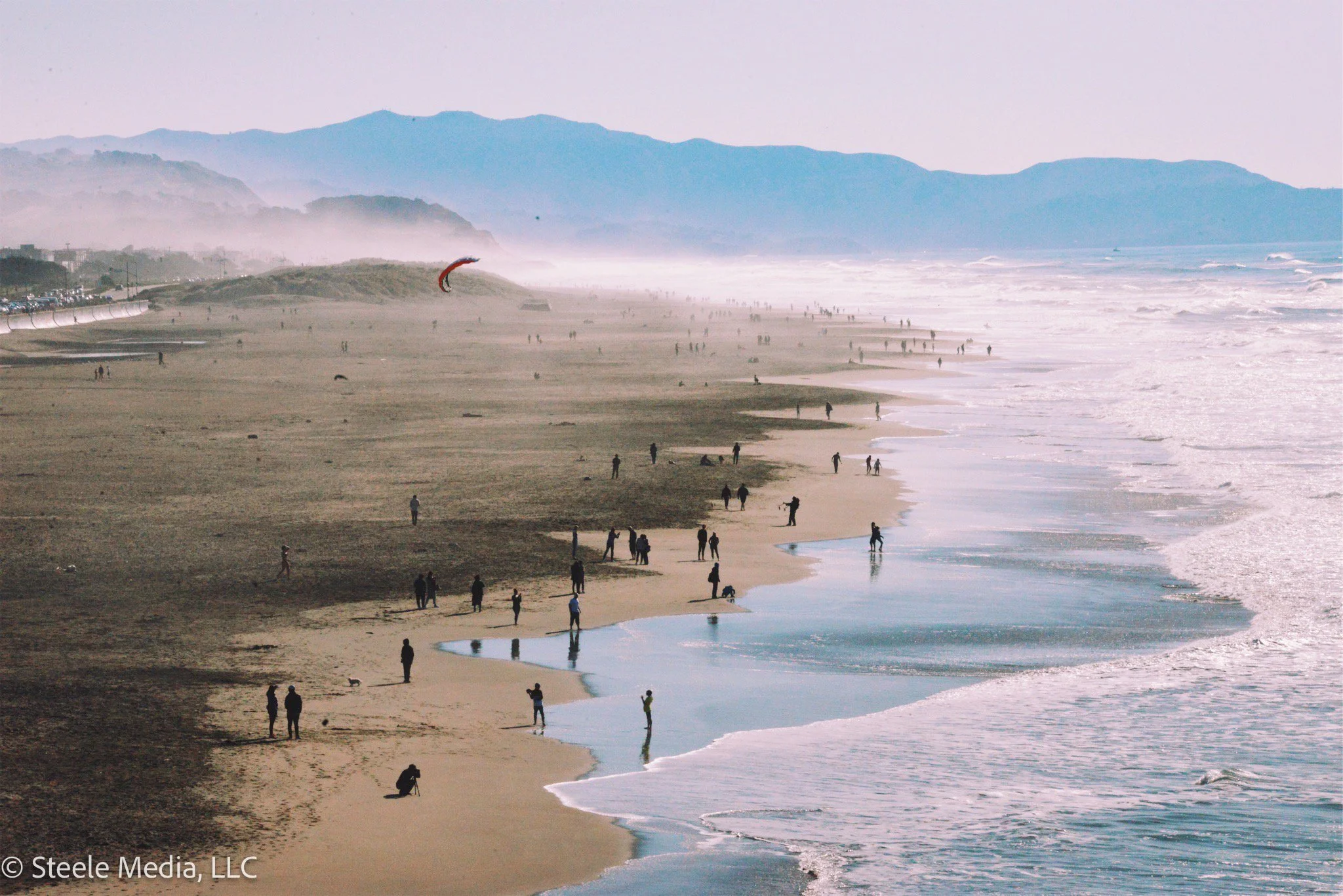 A wide view of a beach with many people walking and standing near the shoreline, some in the water. There are rolling hills and mountains in the background, with a light mist. A kite surfer is visible in the distance.