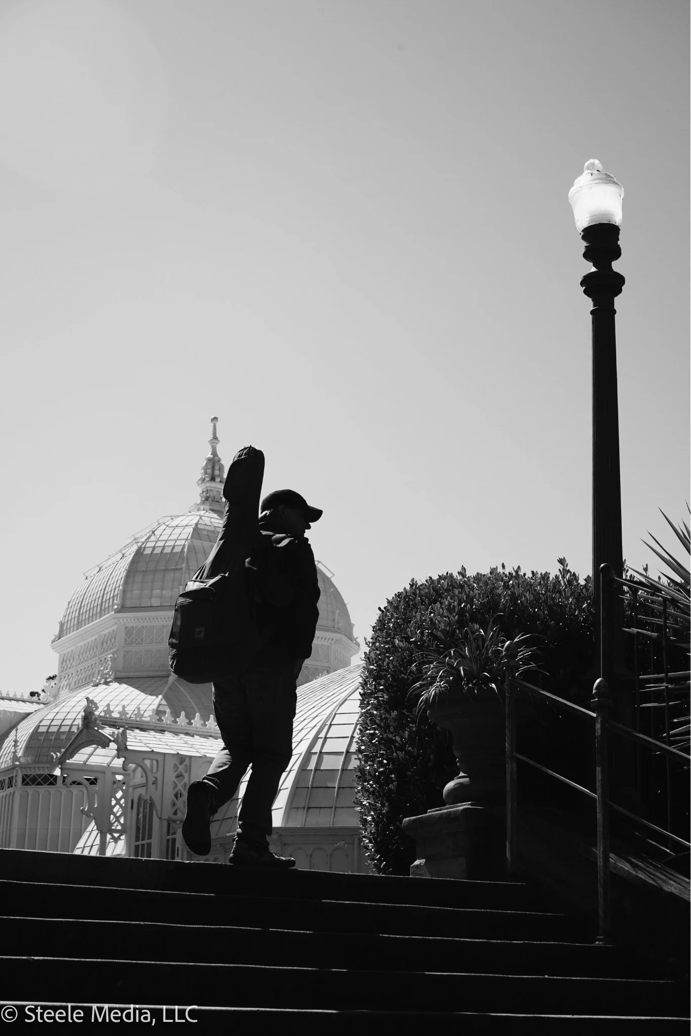 A person with a backpack walking up stairs near a large, ornate glass-domed building, with a streetlamp and bushes in the scene, in black and white.