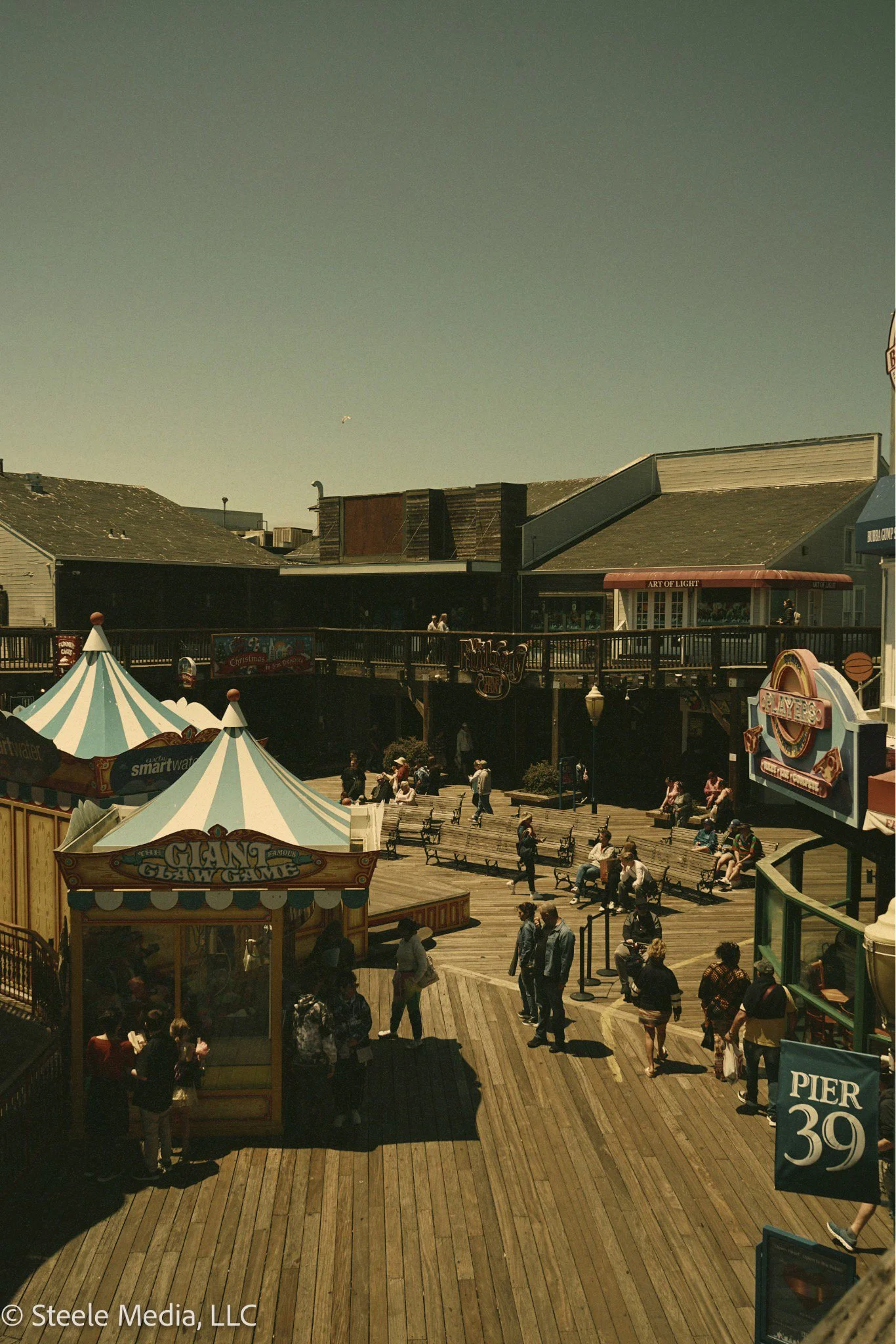 People walking and sitting on benches on a wooden pier with amusement rides and shops, Pier 39 in San Francisco.