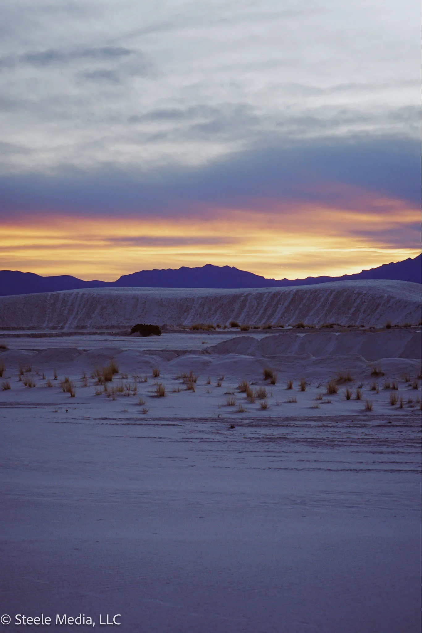 A desert landscape at sunset with mountains in the background, sandy dunes, and sparse vegetation in the foreground.