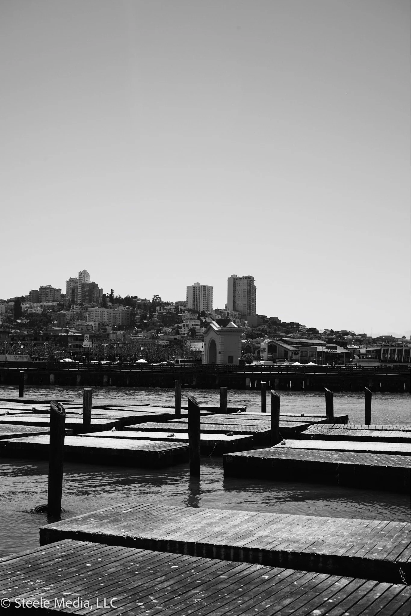 Black and white photo of docks on a waterfront with a city skyline in the background, including tall buildings and a bridge.