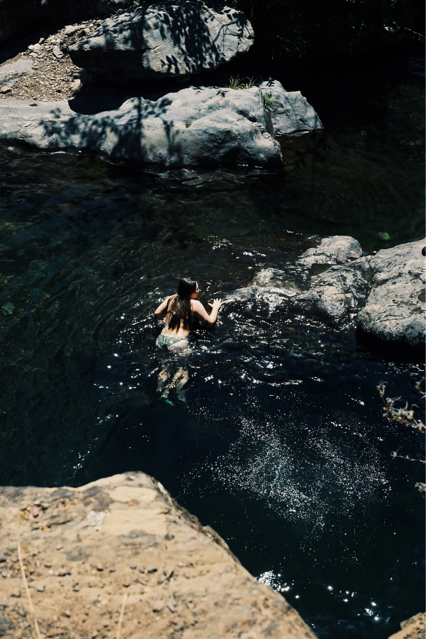 A woman swimming in a rocky river with dark water and large rocks around.