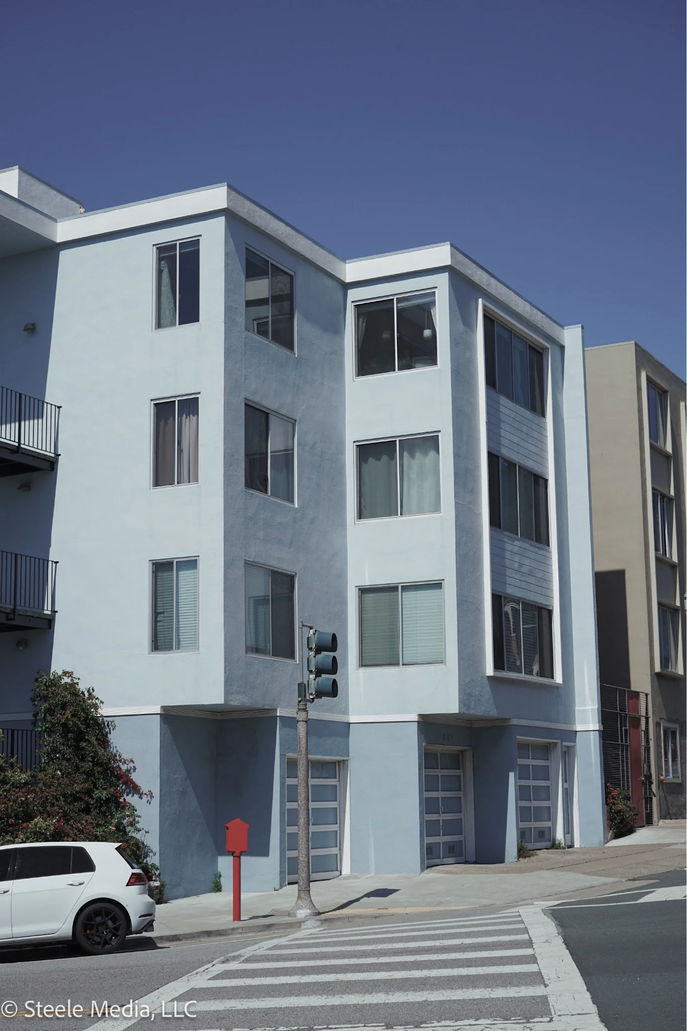 A light blue multi-story residential building with large windows, situated at a street corner with a crosswalk and a white car parked nearby.