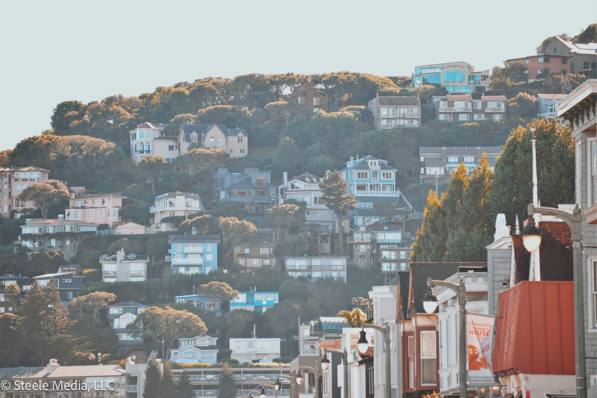 A hillside neighborhood with multiple houses and buildings, and a street with lamp posts and storefronts in the foreground.