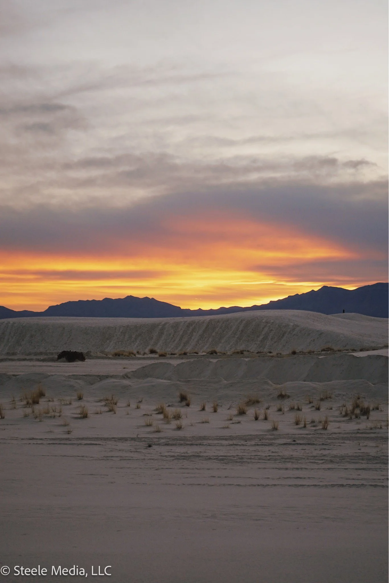 Sunset over a desert landscape with sand dunes, sparse vegetation, and distant mountains under a partly cloudy sky.
