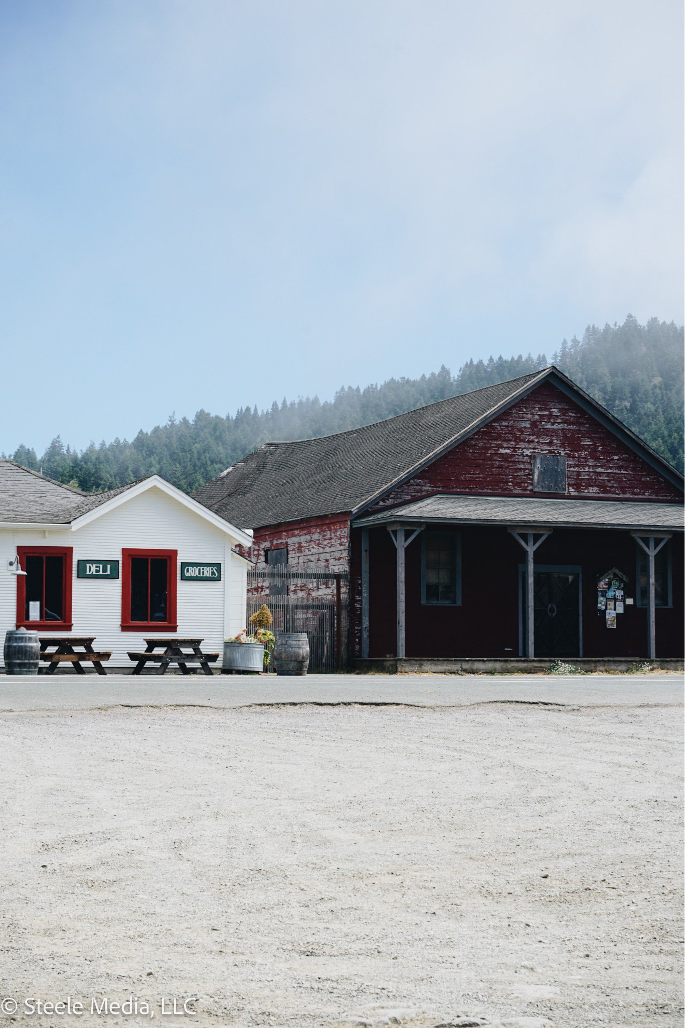 A small white building with red window frames and signs that read 'Deli' and 'Groceries' is situated next to a larger, older red wooden building with a porch. There are picnic tables and barrels in front of the white building, with mountains and a cl