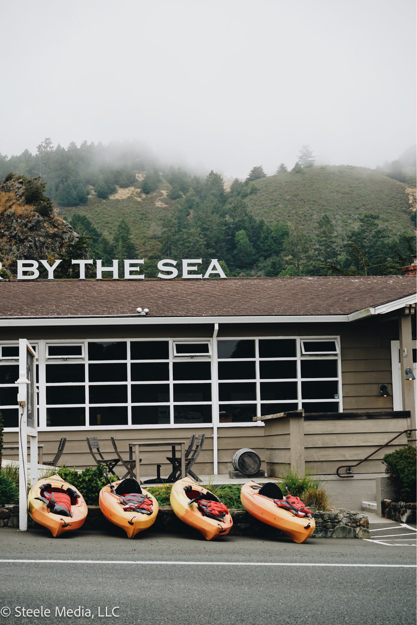 Four kayaks with life jackets and paddles lined up in front of a building with a sign that reads 'BY THE SEA', situated near a parking lot with mountains and fog in the background.