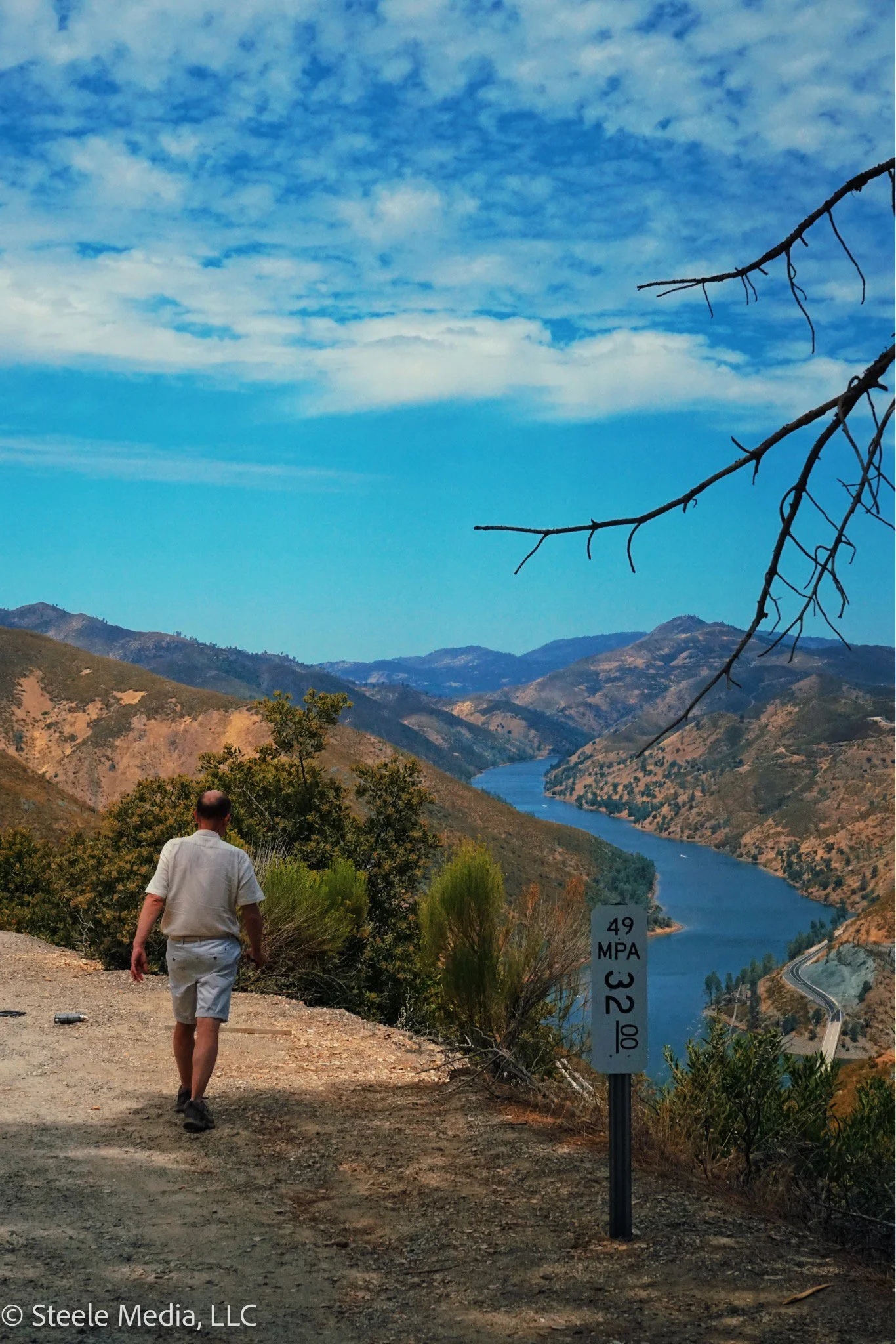 A man walking on a dirt path beside a river in a mountainous landscape, with a sign showing an elevation of 4929 feet and a temperature of 32°F, under a partly cloudy sky.