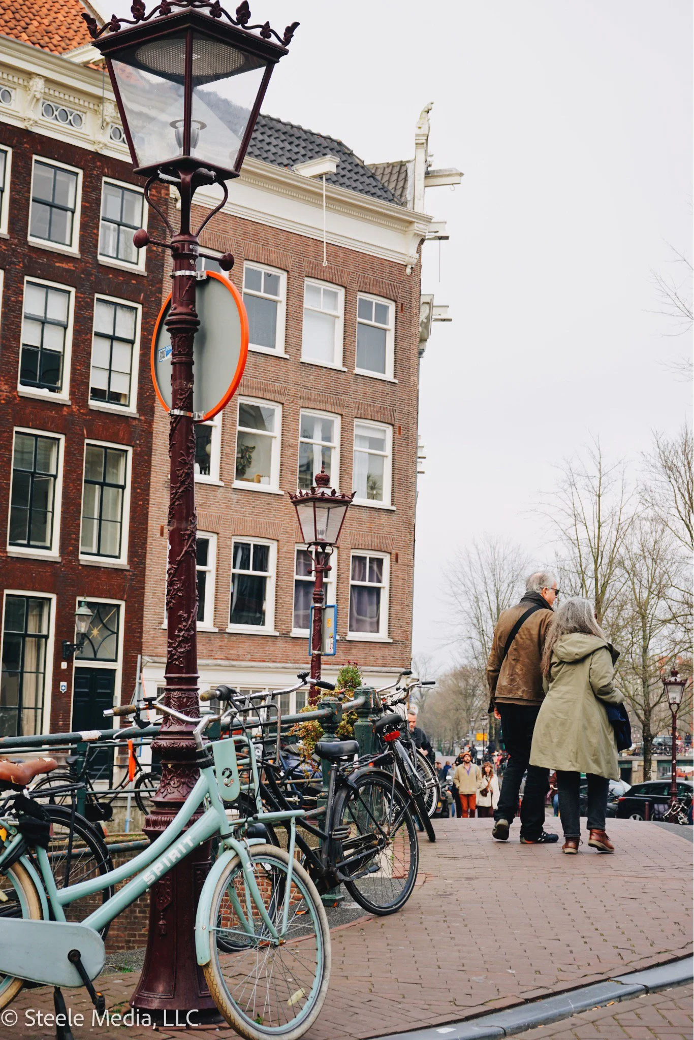 Street scene with vintage street lamps, parked bicycles, and a group of people walking along a sidewalk in front of traditional brick buildings with large windows.