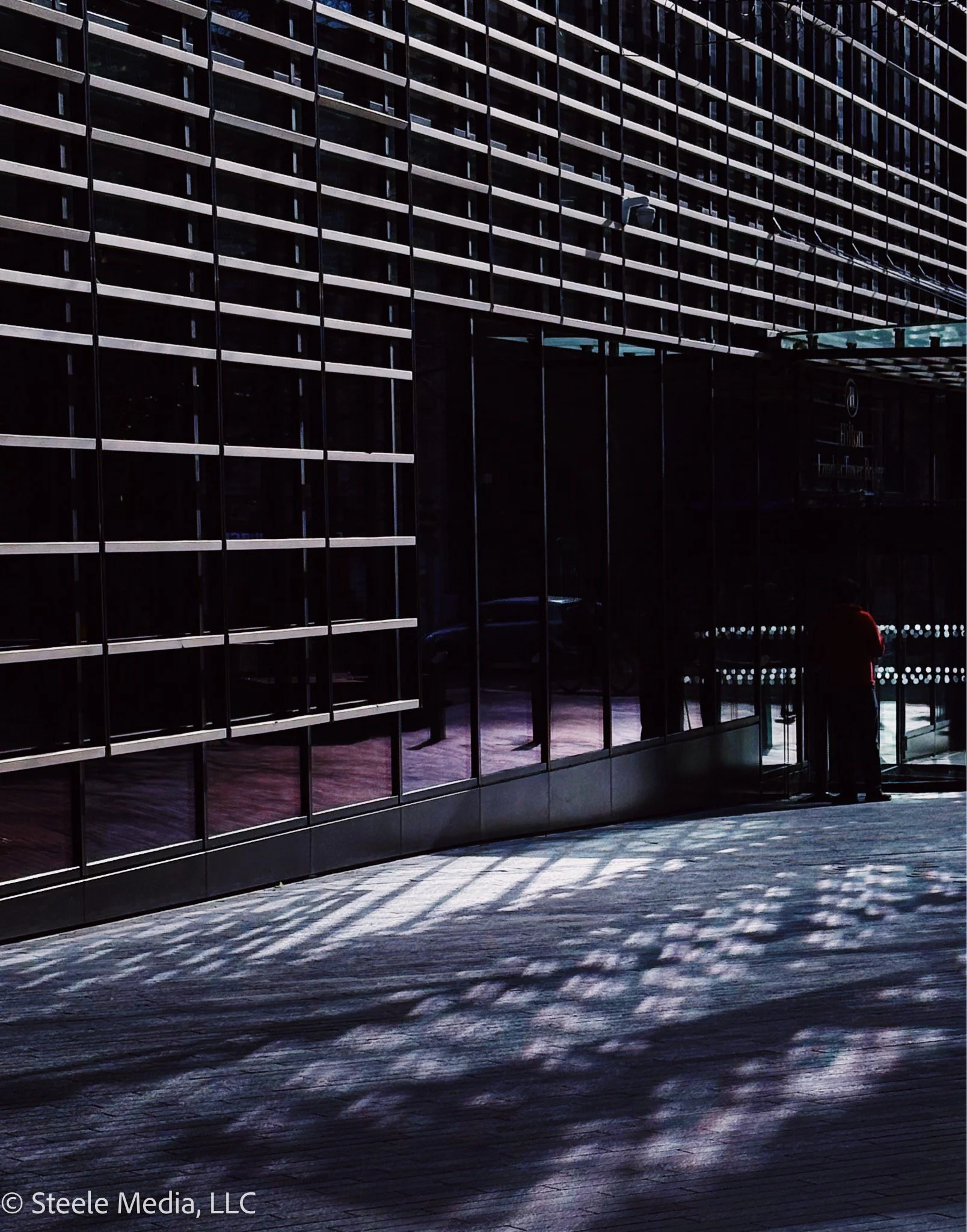 A modern building exterior with large glass windows and horizontal metal slats, with sunlight casting shadow patterns on the sidewalk, and a person standing near the entrance.