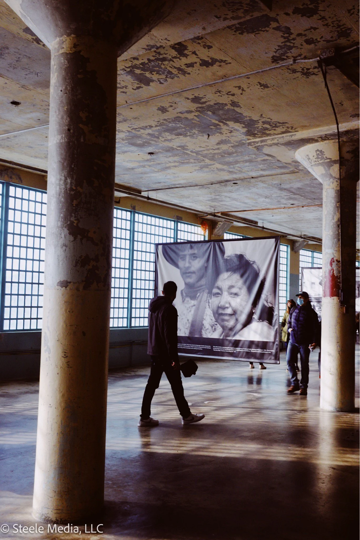 People walking inside an industrial-style building with large windows, viewing a large black-and-white photograph of two people on display.