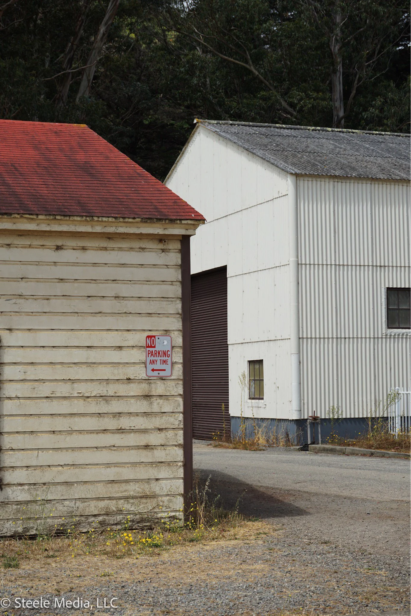 Photo of two buildings, one with a red-tiled roof and the other with a grey, corrugated metal roof, with a 'No Parking Any Time' sign on the beige wooden building in the foreground.