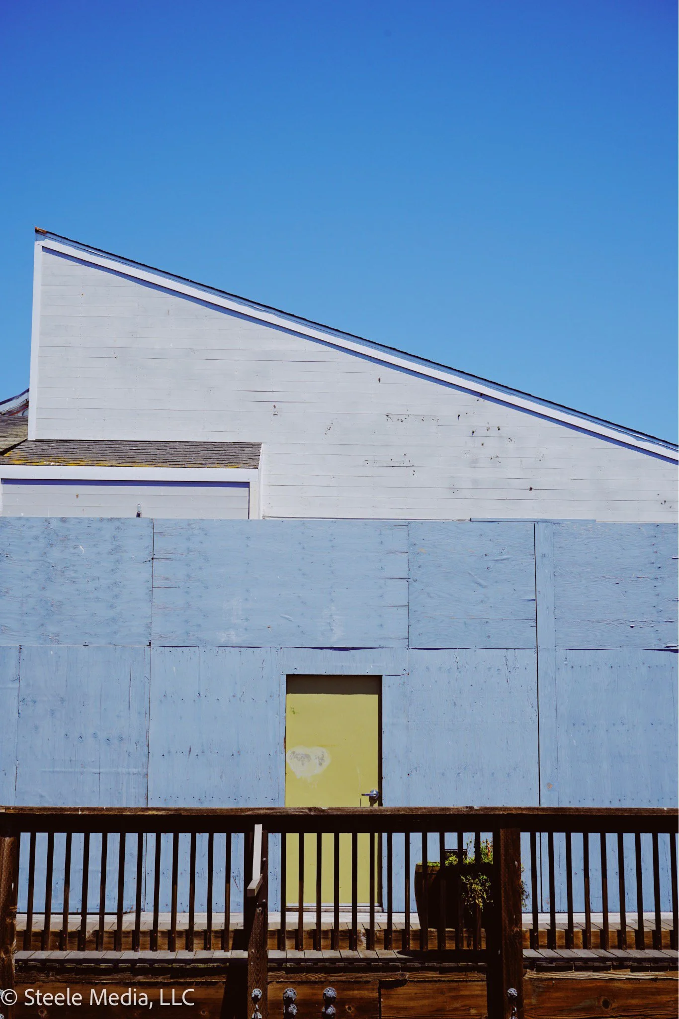 A white building with a sloped roof and small side extension, covered by a wooden fence and some potted plants, against a clear blue sky.