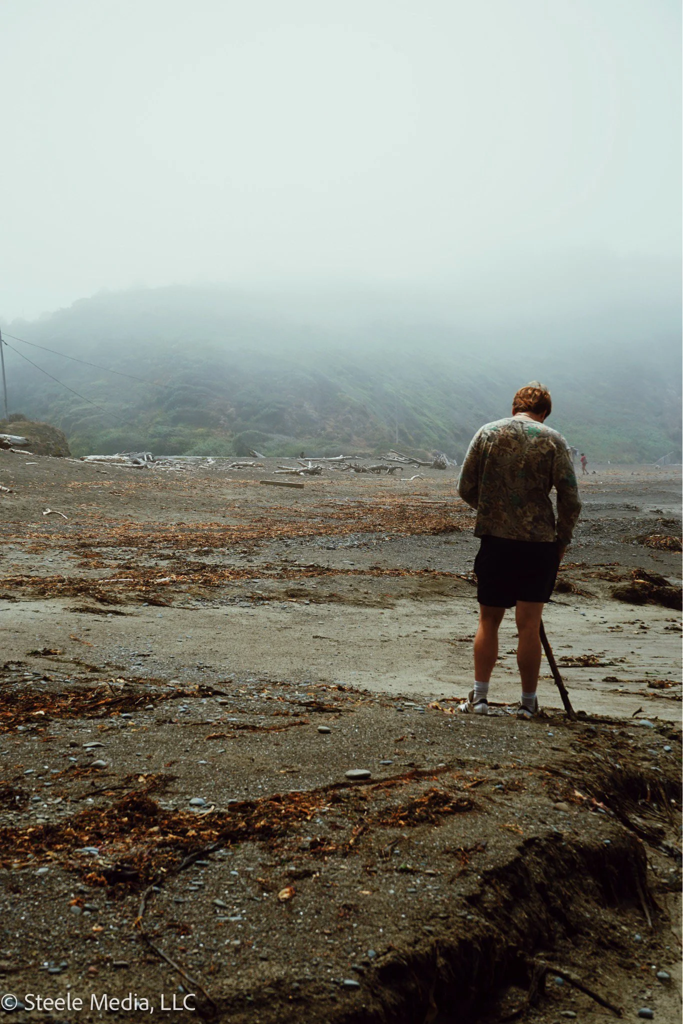 Person with short hair wearing a camouflage jacket and shorts, standing on a foggy beach or coastal area, looking down at the ground while holding a stick or tool.