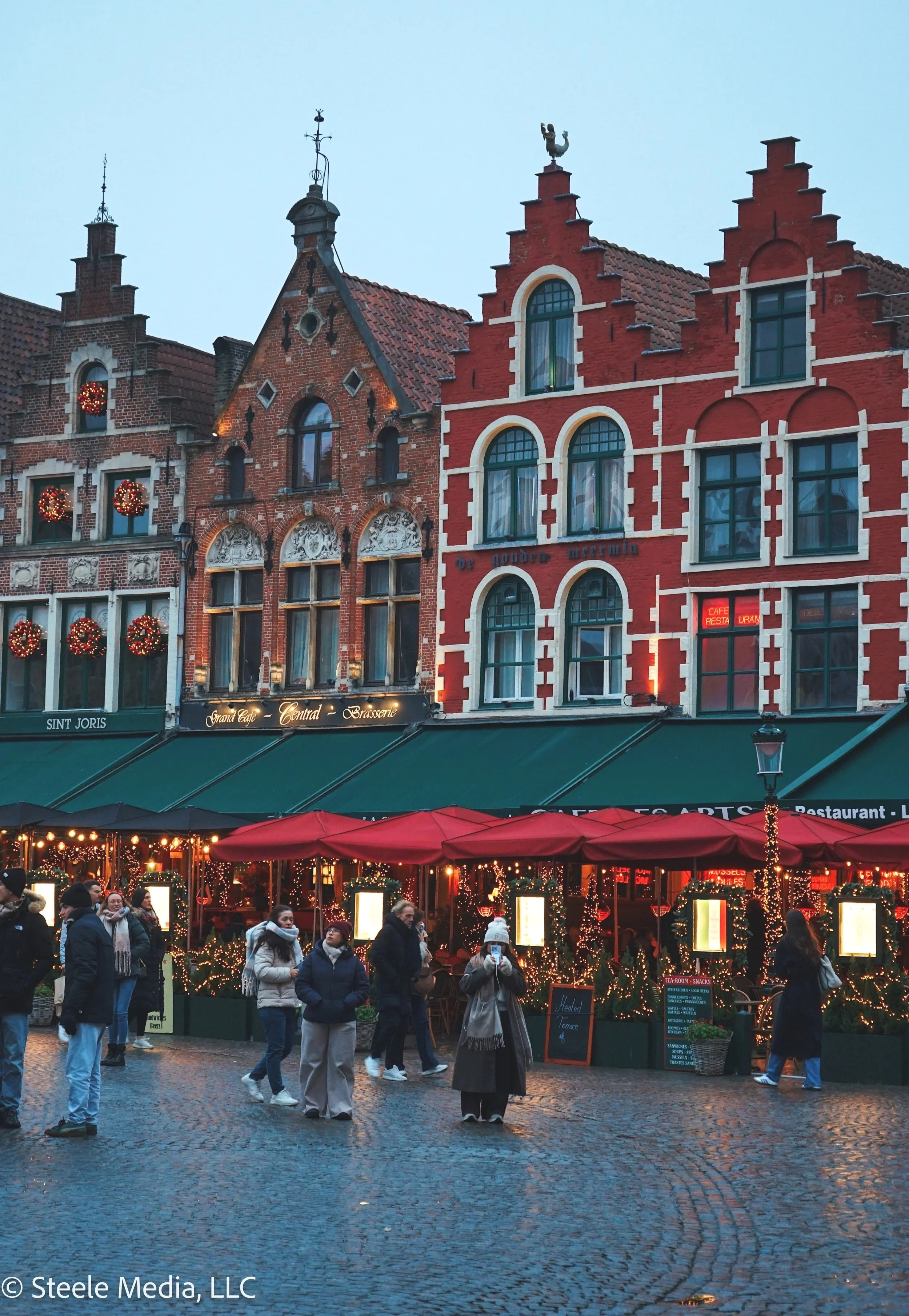 A European-style town square at dusk decorated for Christmas, with historic buildings and people walking past a festive outdoor market with red umbrellas and twinkling lights.