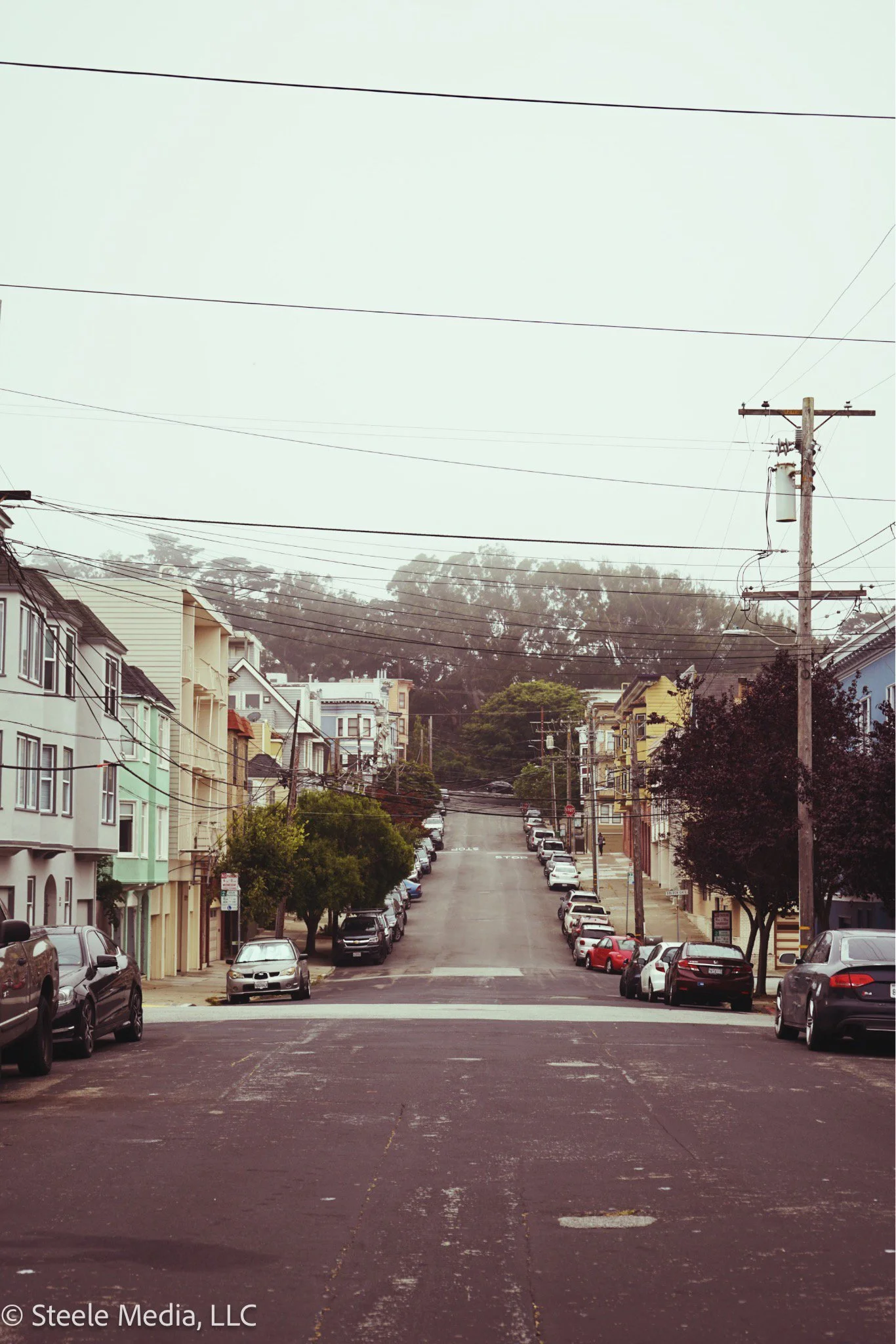 A city street with parked cars along both sides, colorful residential buildings, trees, utility poles, and power lines under a cloudy sky.
