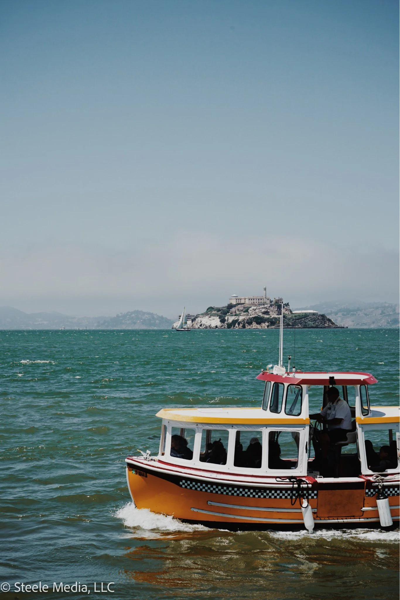 A yellow and white boat on the water near Alcatraz Island with a sailboat in the distance.