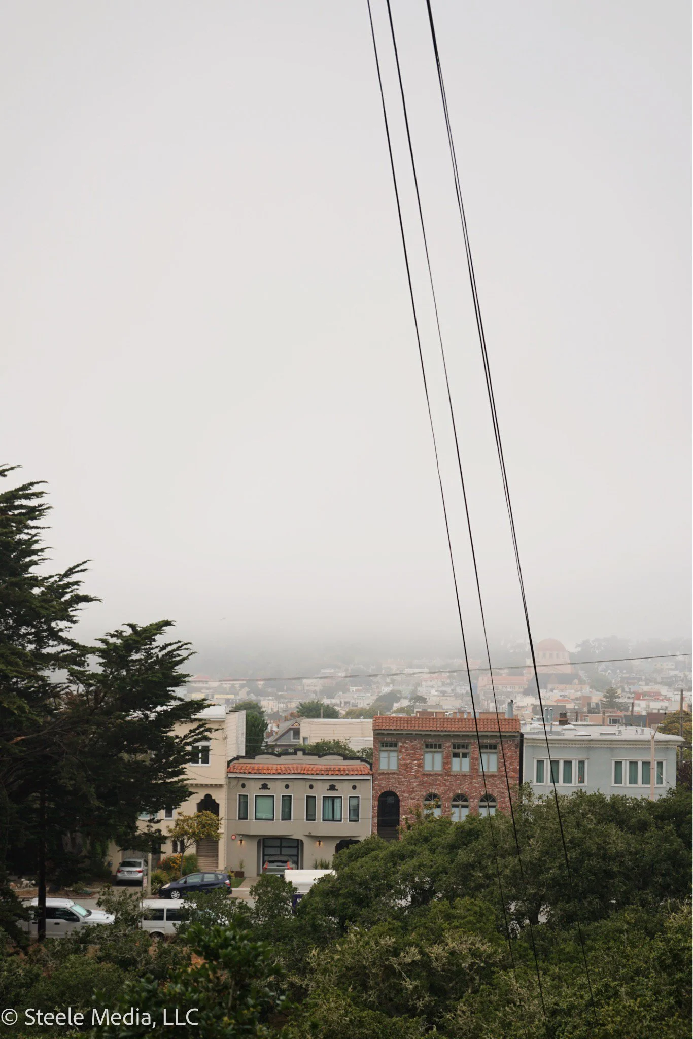 A foggy cityscape with residential buildings, trees in the foreground, and power lines overhead on a cloudy day.
