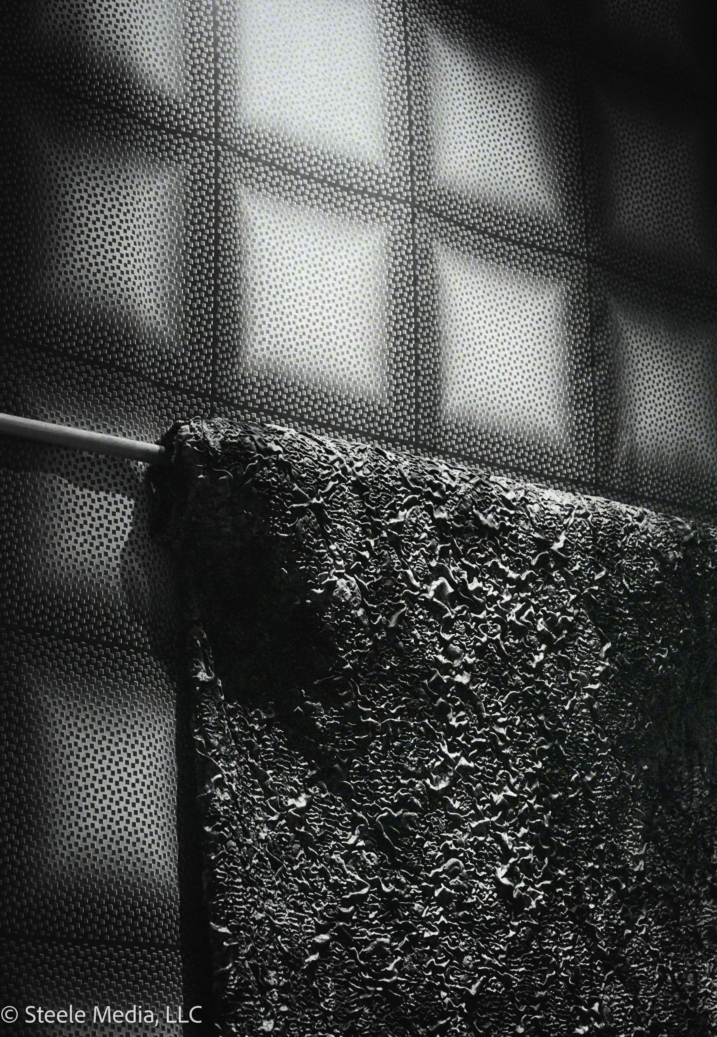 Close-up of a black irregularly textured object on a metallic surface with a grid pattern, with out-of-focus bright office windows in the background.