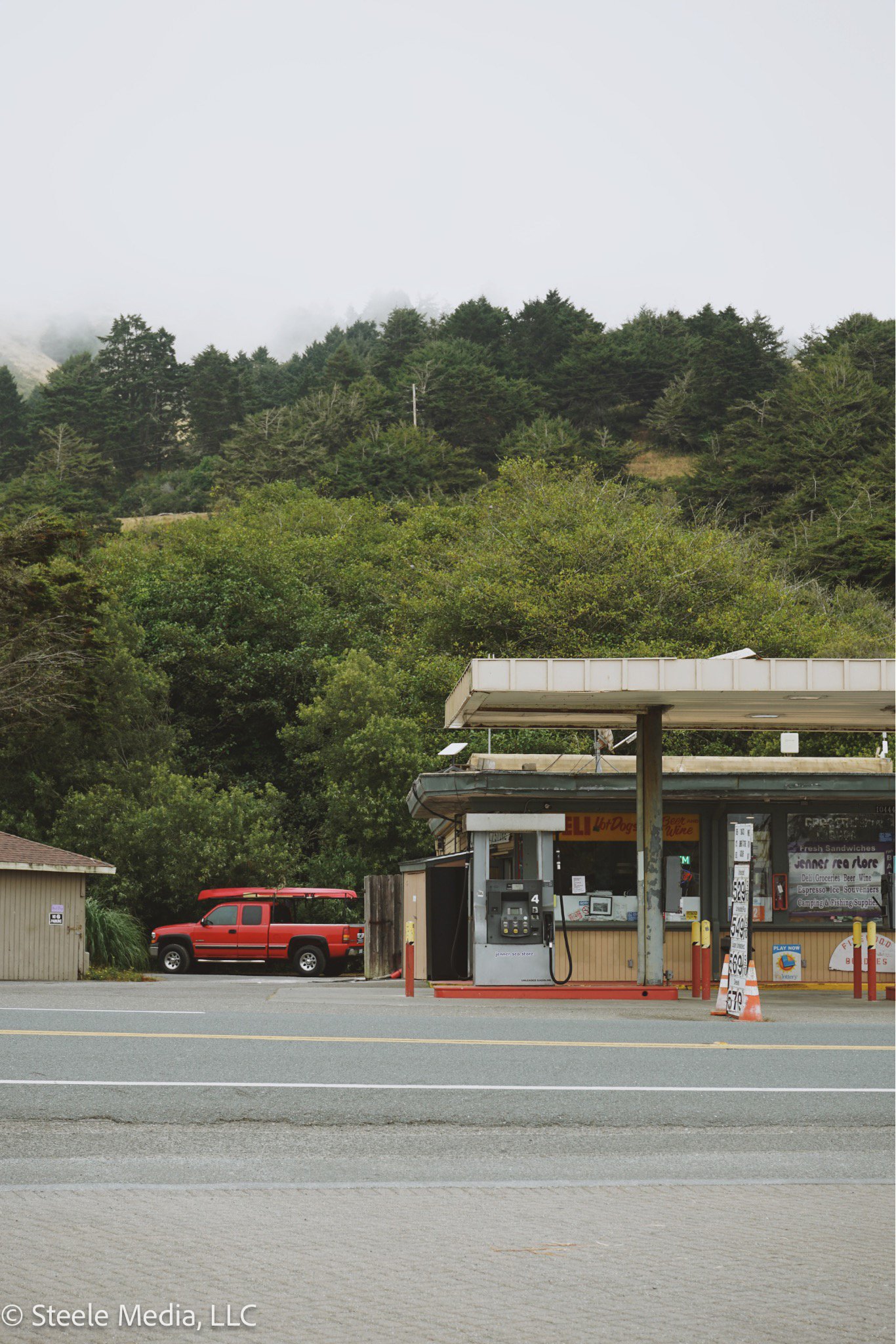 A small roadside convenience store with a gas pump in front, situated at the edge of a forested area with hills and fog in the background.