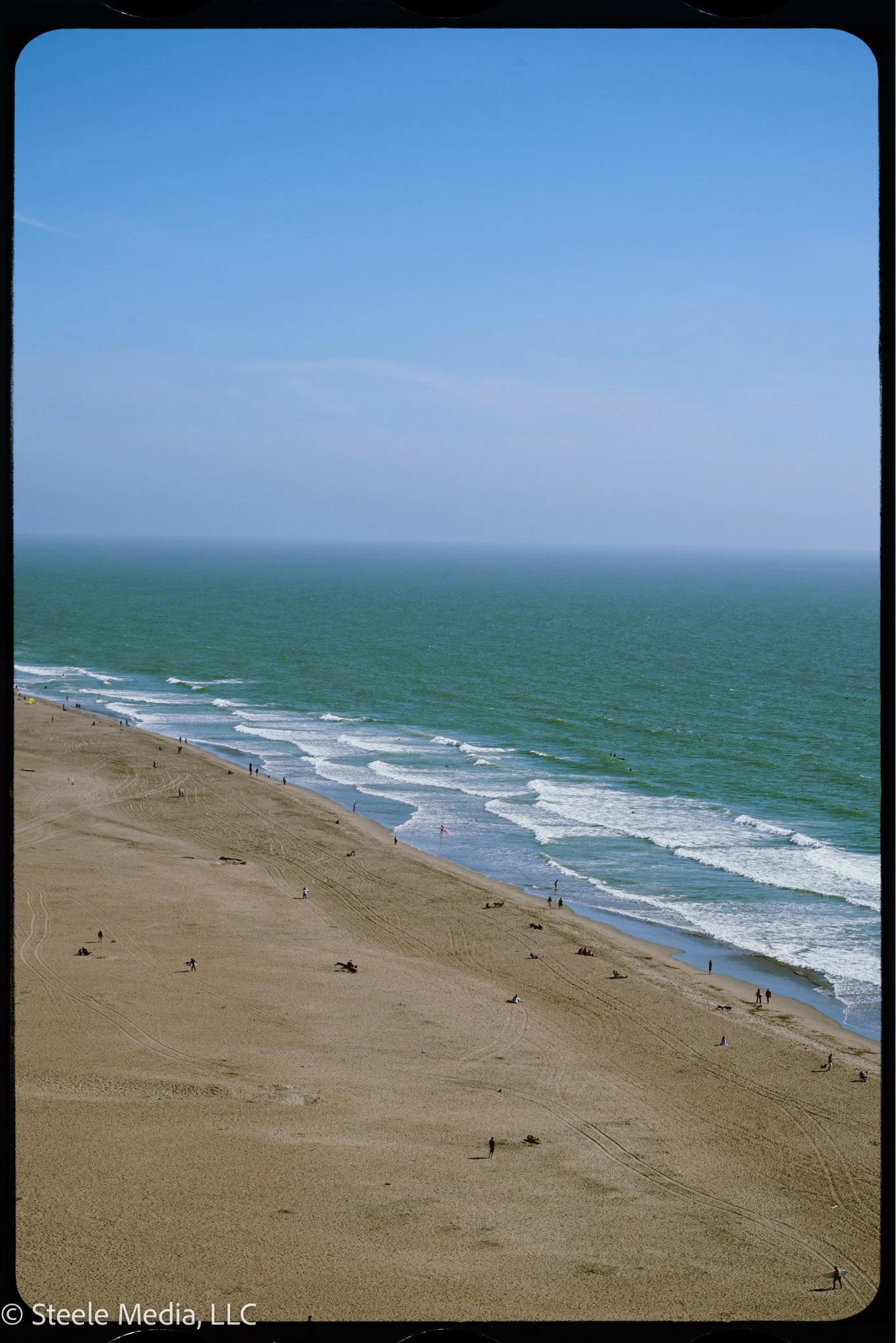 A wide view of a sandy beach with a few people walking and relaxing, gentle waves crashing onto the shore, and a clear blue sky.
