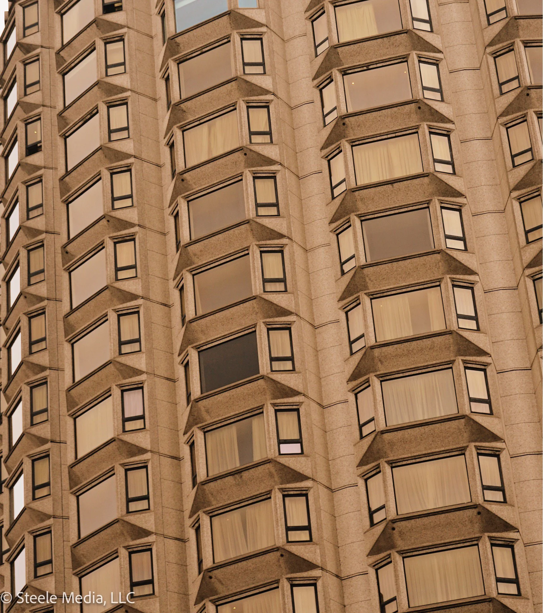 Close-up view of a tall building with numerous bay windows in a grid pattern, with some windows having curtains and others appearing dark.
