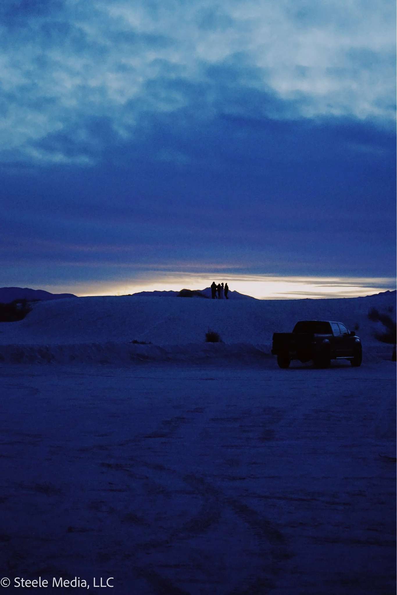 Silhouetted group of people standing on a sand dune at sunset or sunrise, with a dark vehicle parked nearby, under a sky with layered clouds in blue and purple hues.
