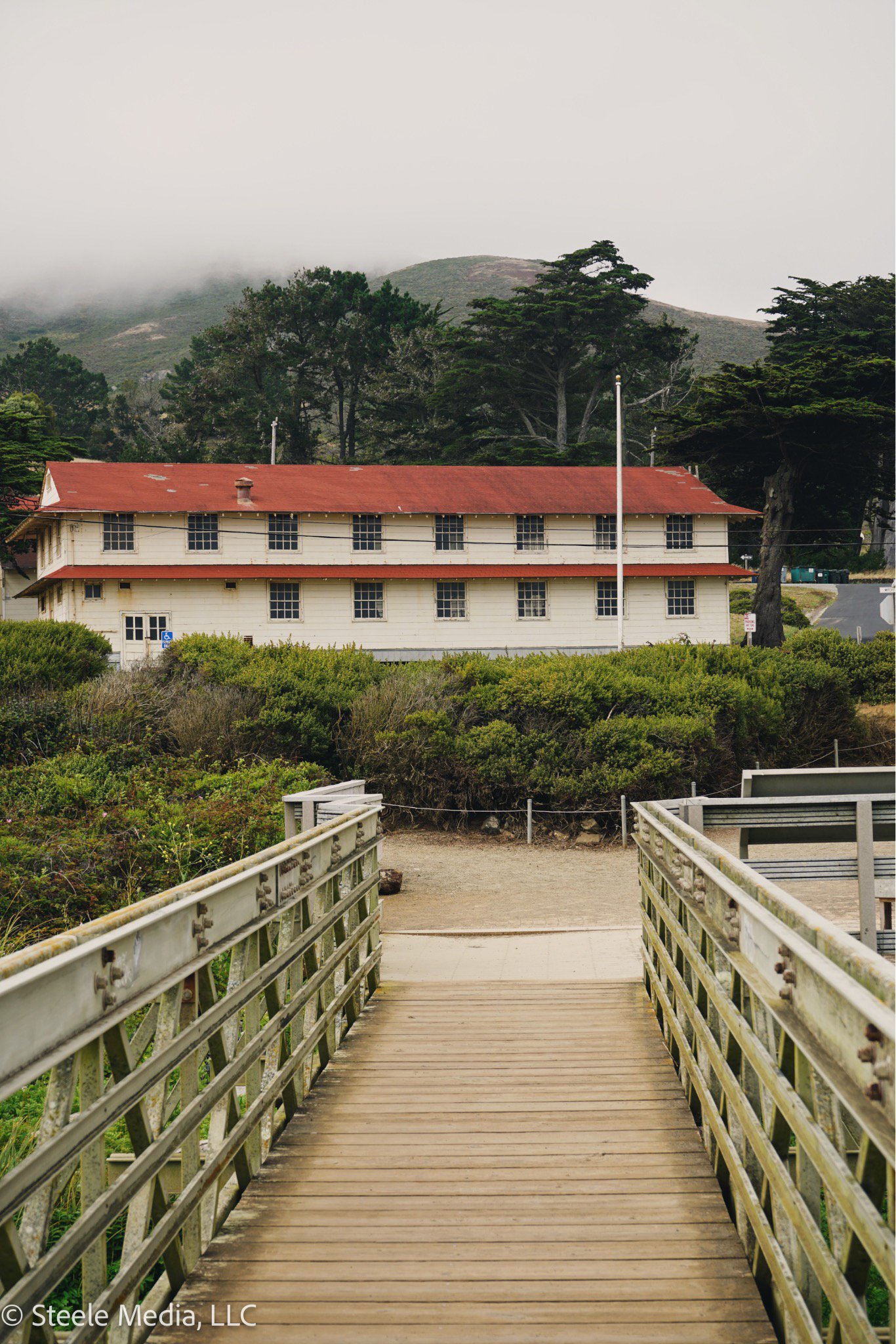 A wooden pathway leading to a large two-story house with a red roof, surrounded by green bushes and trees, with foggy mountains in the background.