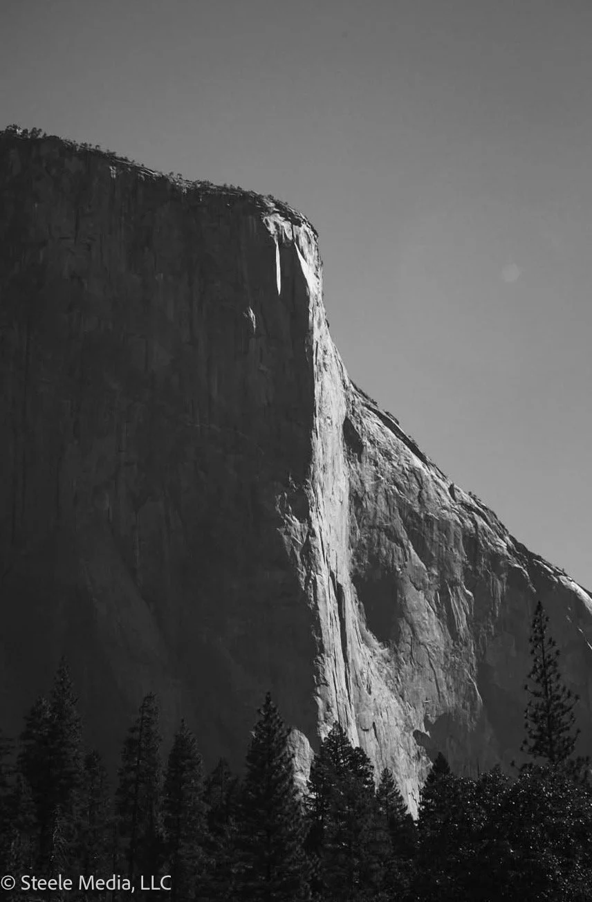 Black and white photograph of a large mountain with a steep cliff, trees at the base, and a clear sky above.