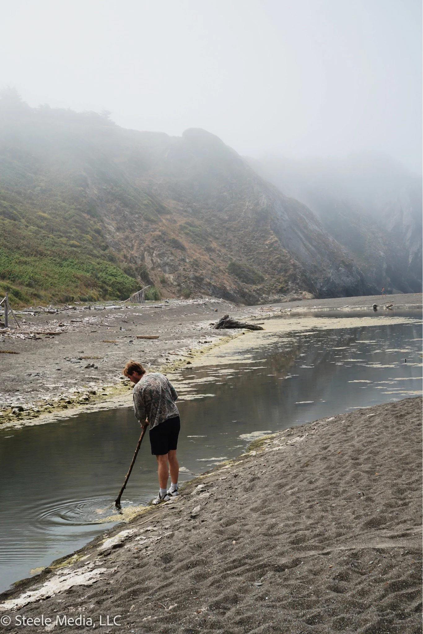 A person wearing black shorts, a patterned jacket, and white shoes stands on a sandy, rocky beach near a body of water, holding a stick and appears to be raking or stirring the water. The scene is misty, with fog hovering over cliffs and mountains in