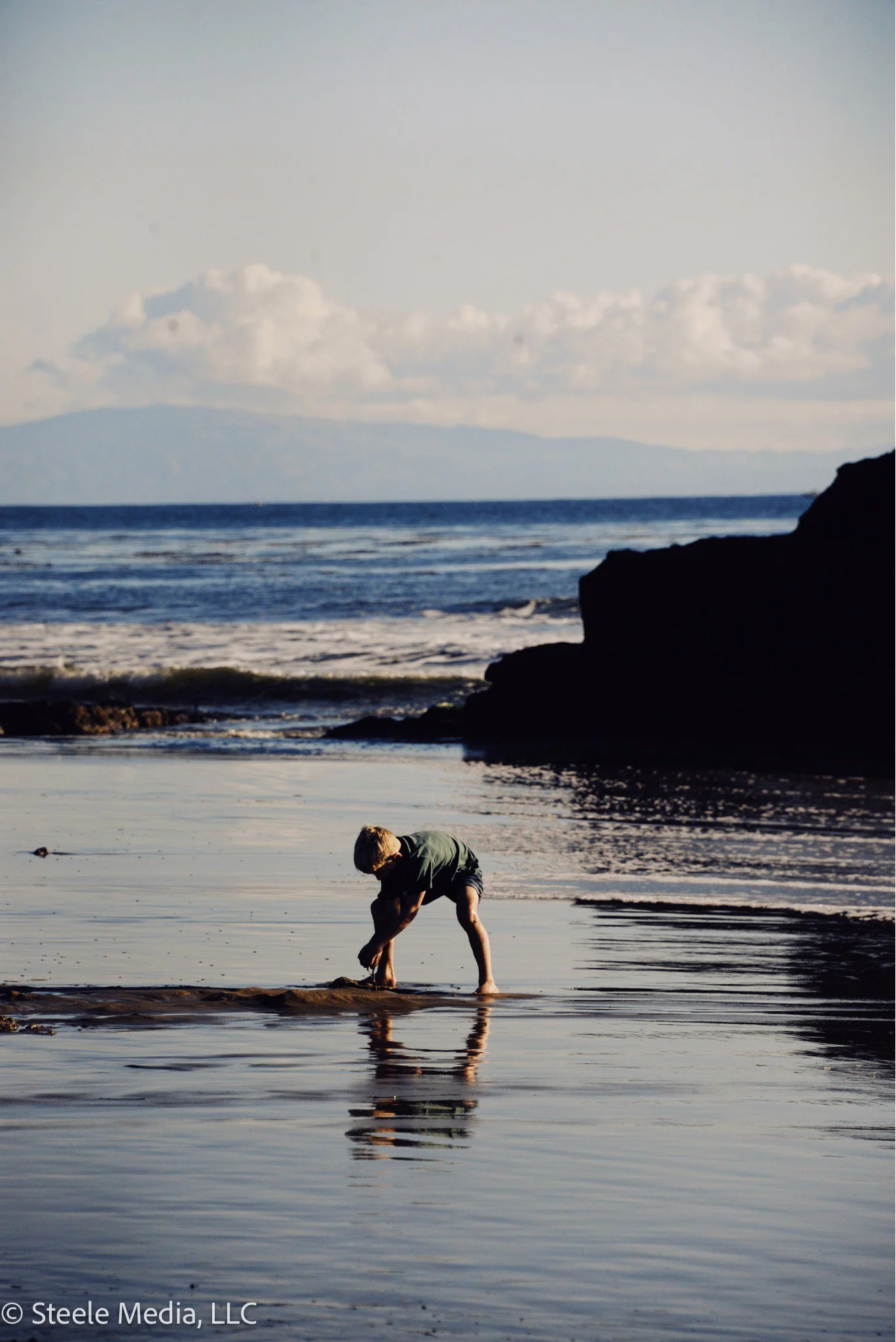 A young boy in a green shirt and shorts playing on a sandy beach near the water, with ocean waves and a distant landmass or mountain in the background.