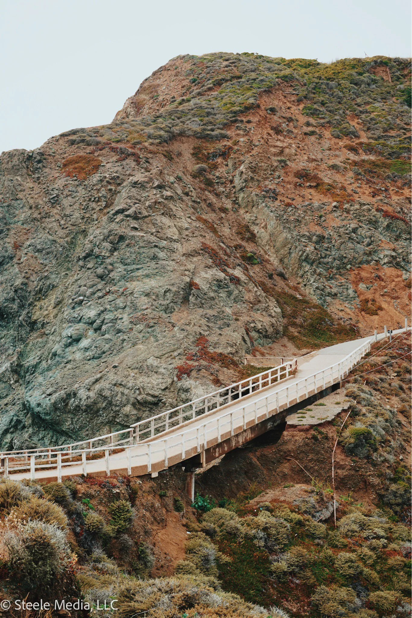 A winding white pedestrian bridge extending through a rugged, rocky hillside with sparse vegetation.