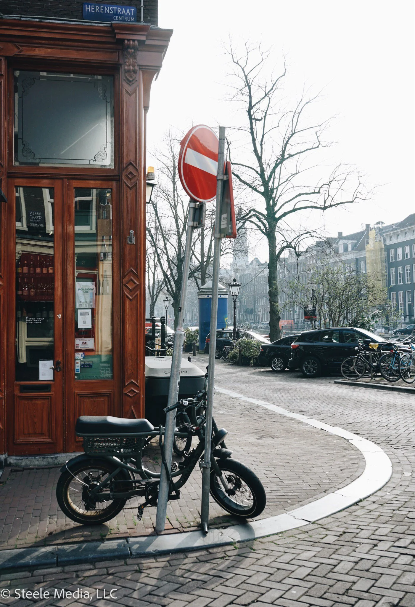 A street scene in a European city with cobblestone sidewalk, a small black scooter parked next to a no-entry traffic sign, a tree with no leaves, a wooden building with a glass door, and parked cars and bicycles along the street.