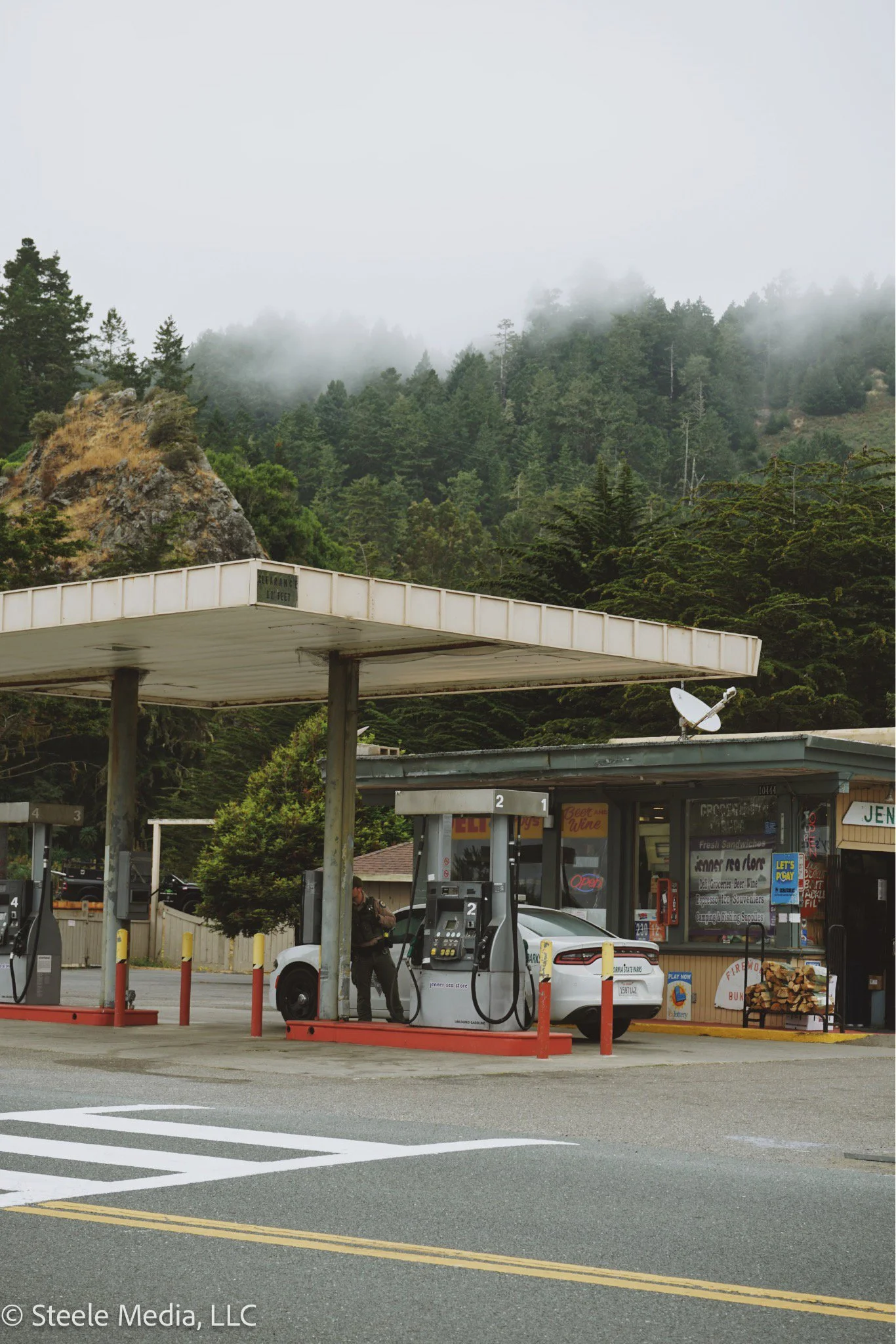 A white car parked at a gas station with a person pumping gas. There are multiple fuel pumps, small shops or kiosks, and a mountainous background with trees and mist.