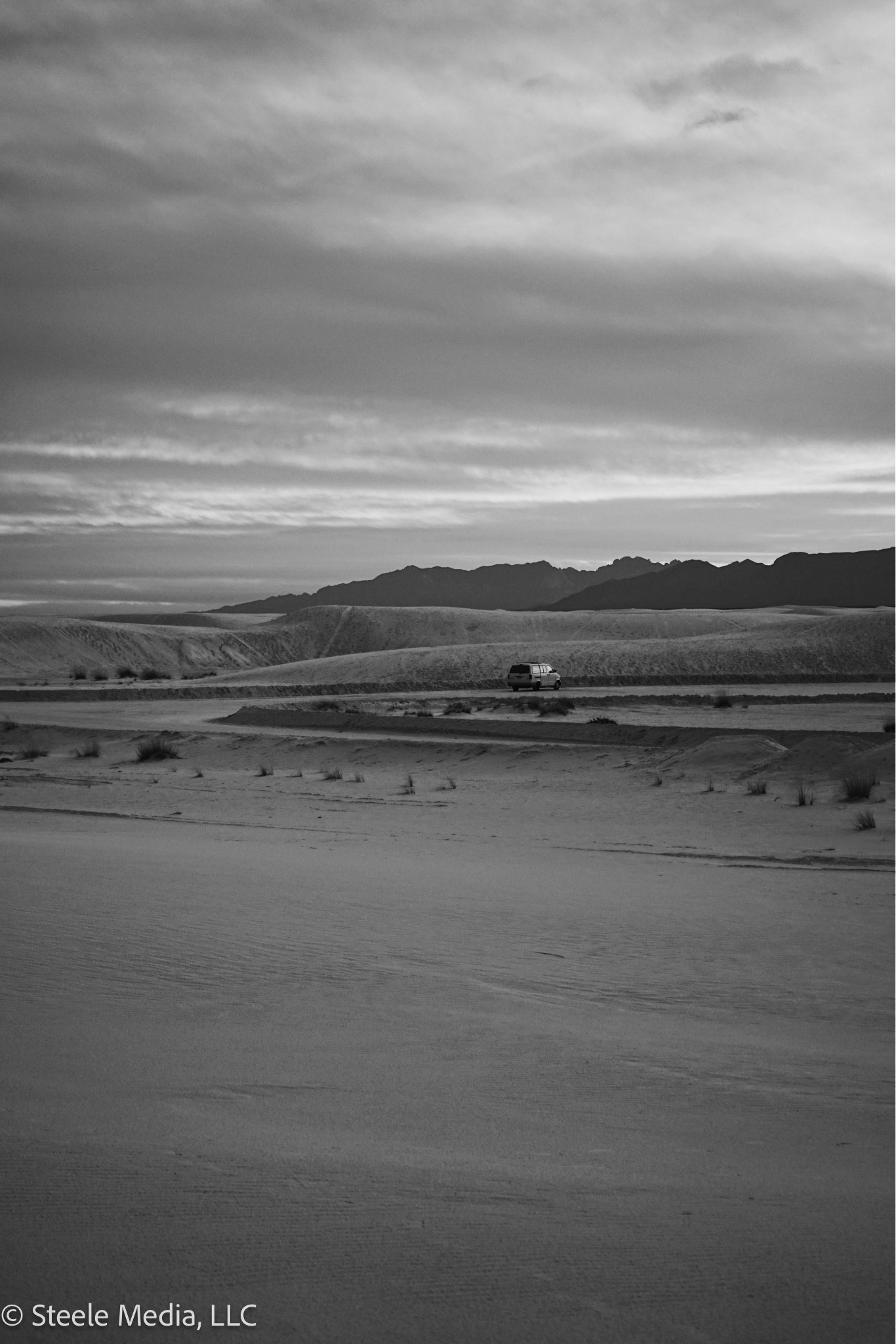 A black and white photo of a desert landscape featuring sand dunes, sparse vegetation, a distant mountain range, and a vehicle traveling on a road.