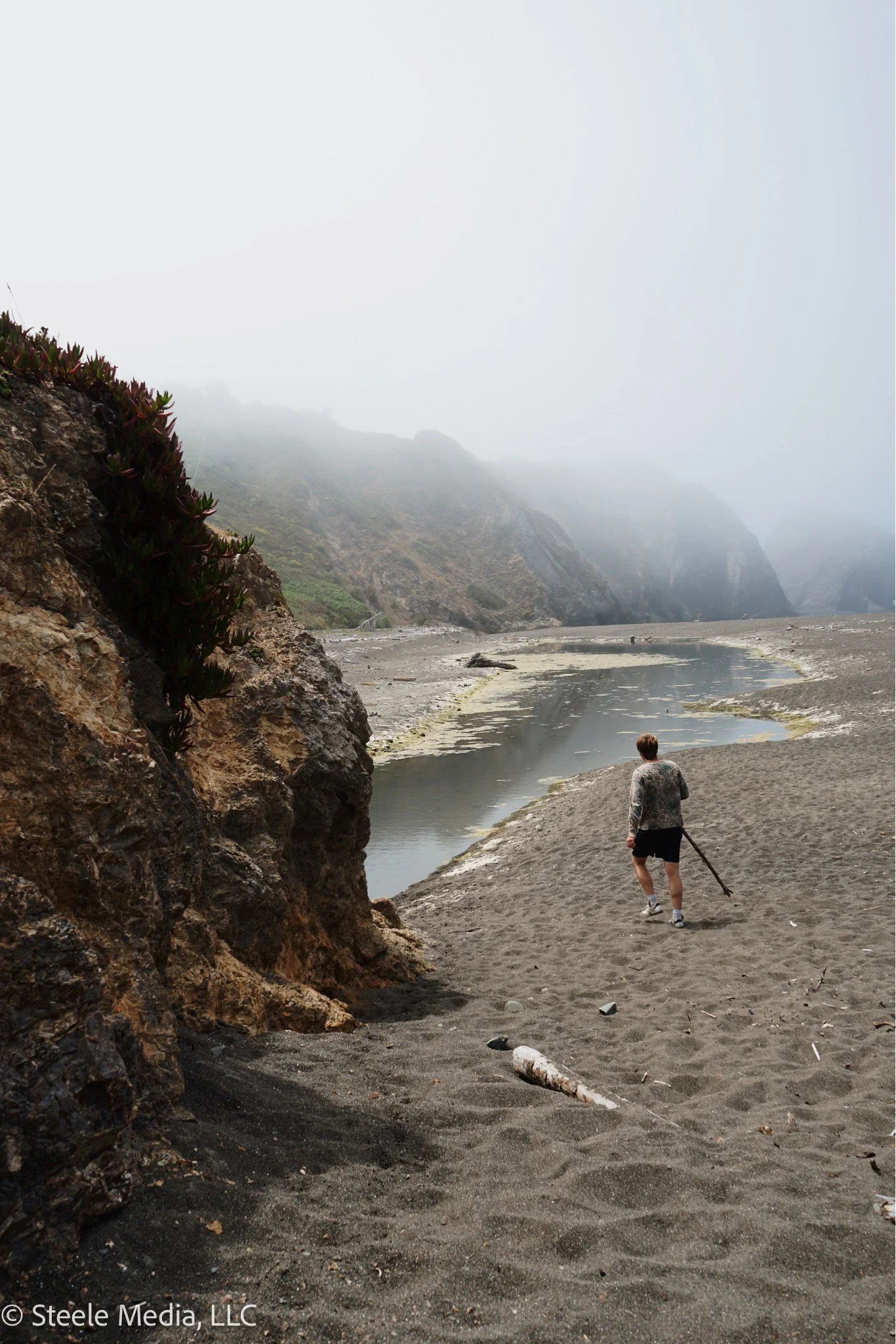 A person walking along a foggy coastal beach with rocky cliffs and a small stream in the background.