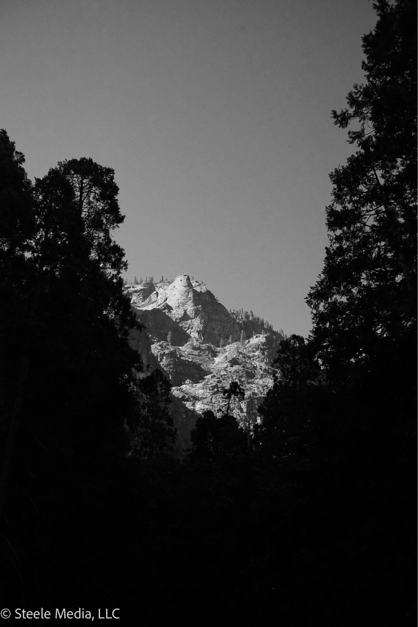 Black and white photo of a mountain with rocky peaks, surrounded by trees, under a clear sky.
