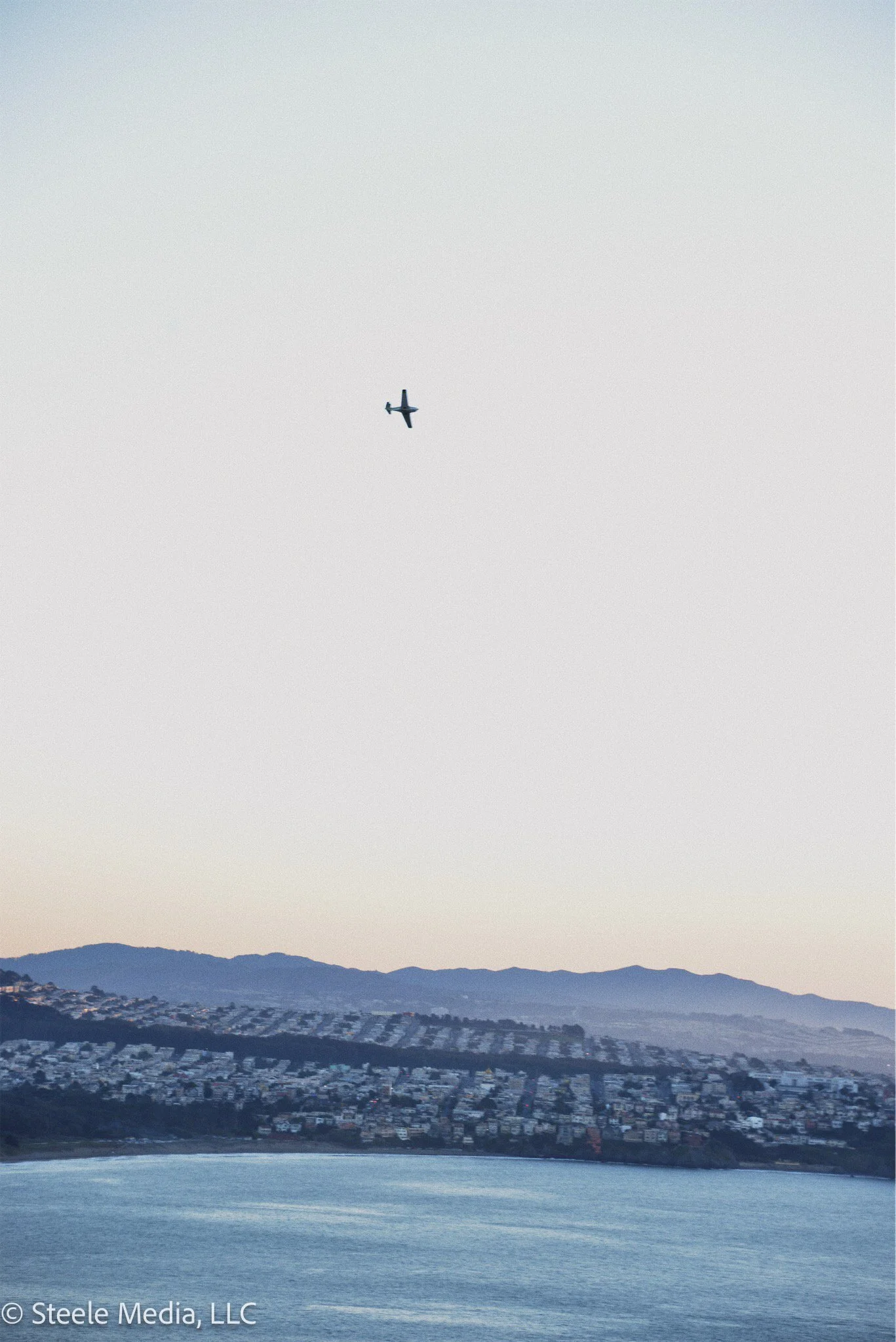 An airplane flying above a body of water near a hilly residential area with houses, with a mountain range in the background.