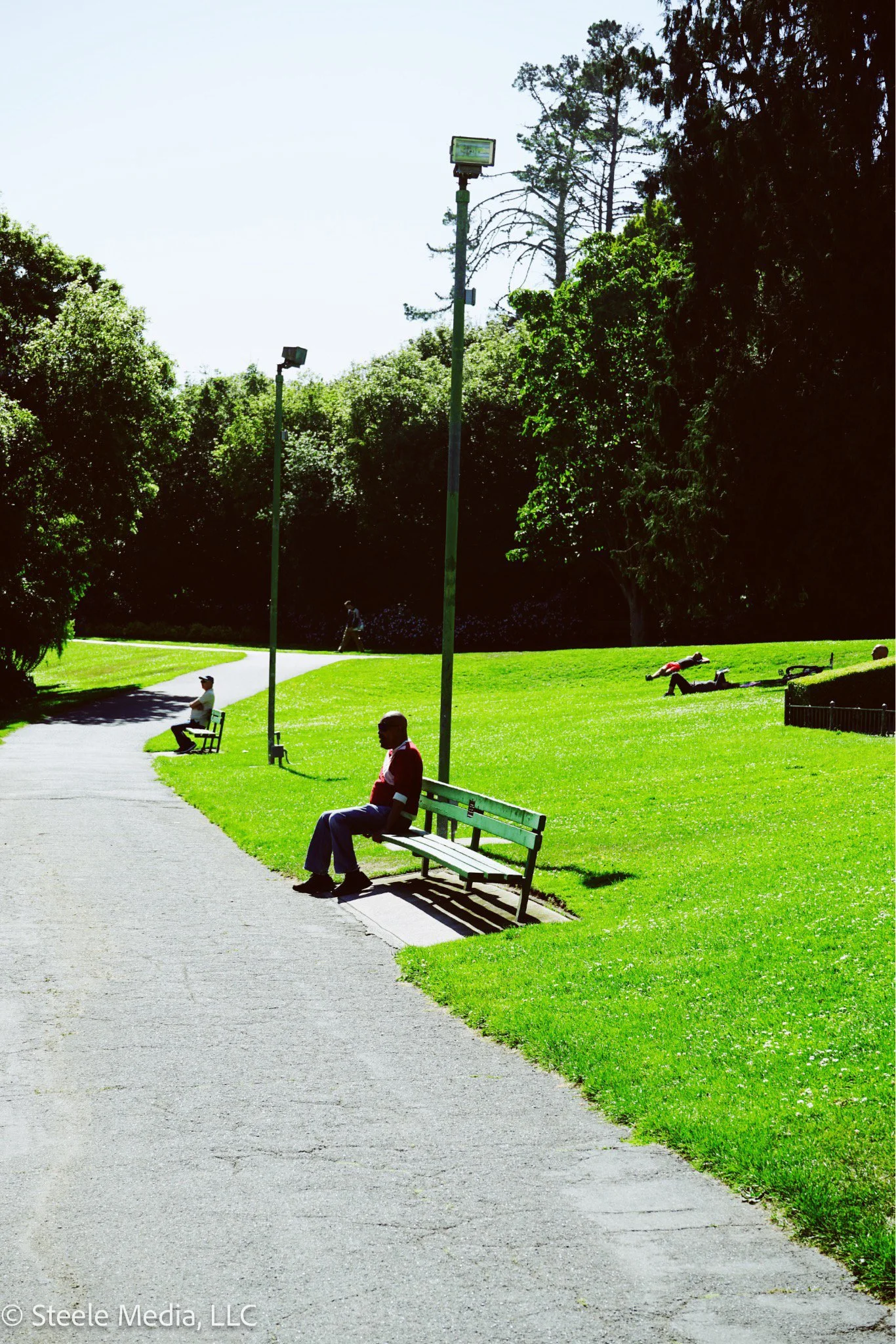 People sitting on benches in a park with a grassy lawn, trees, and streetlights, on a sunny day.