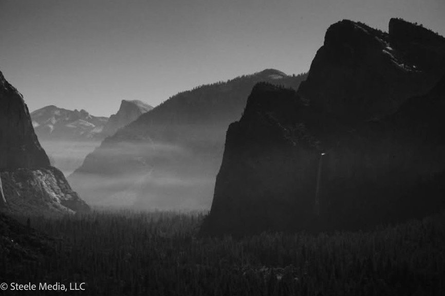 Black and white landscape of mountains and a valley with trees, with mist and clouds in the background.