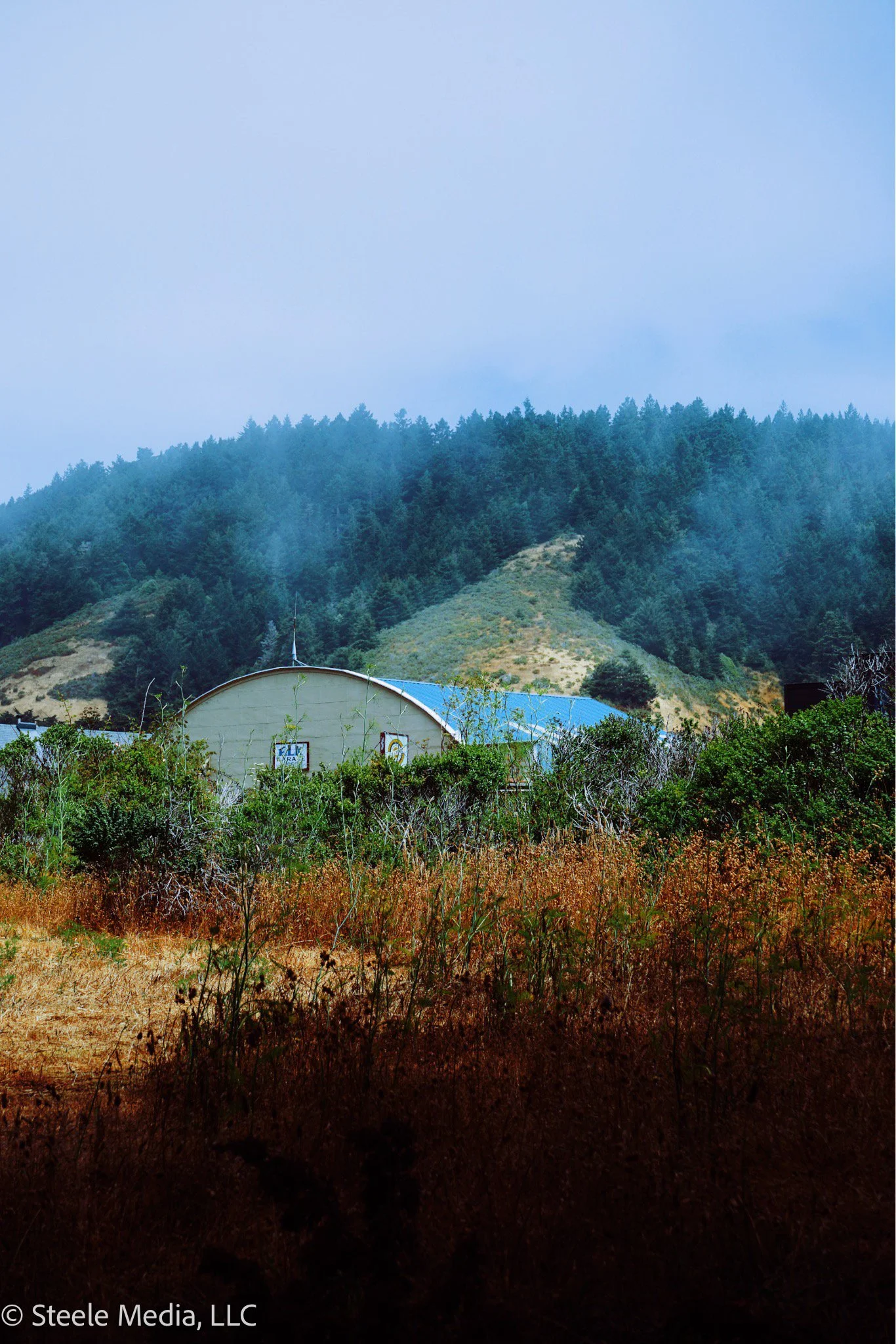 Rural scene with a domed building surrounded by shrubs and grass, forested hills in the background under a cloudy sky.