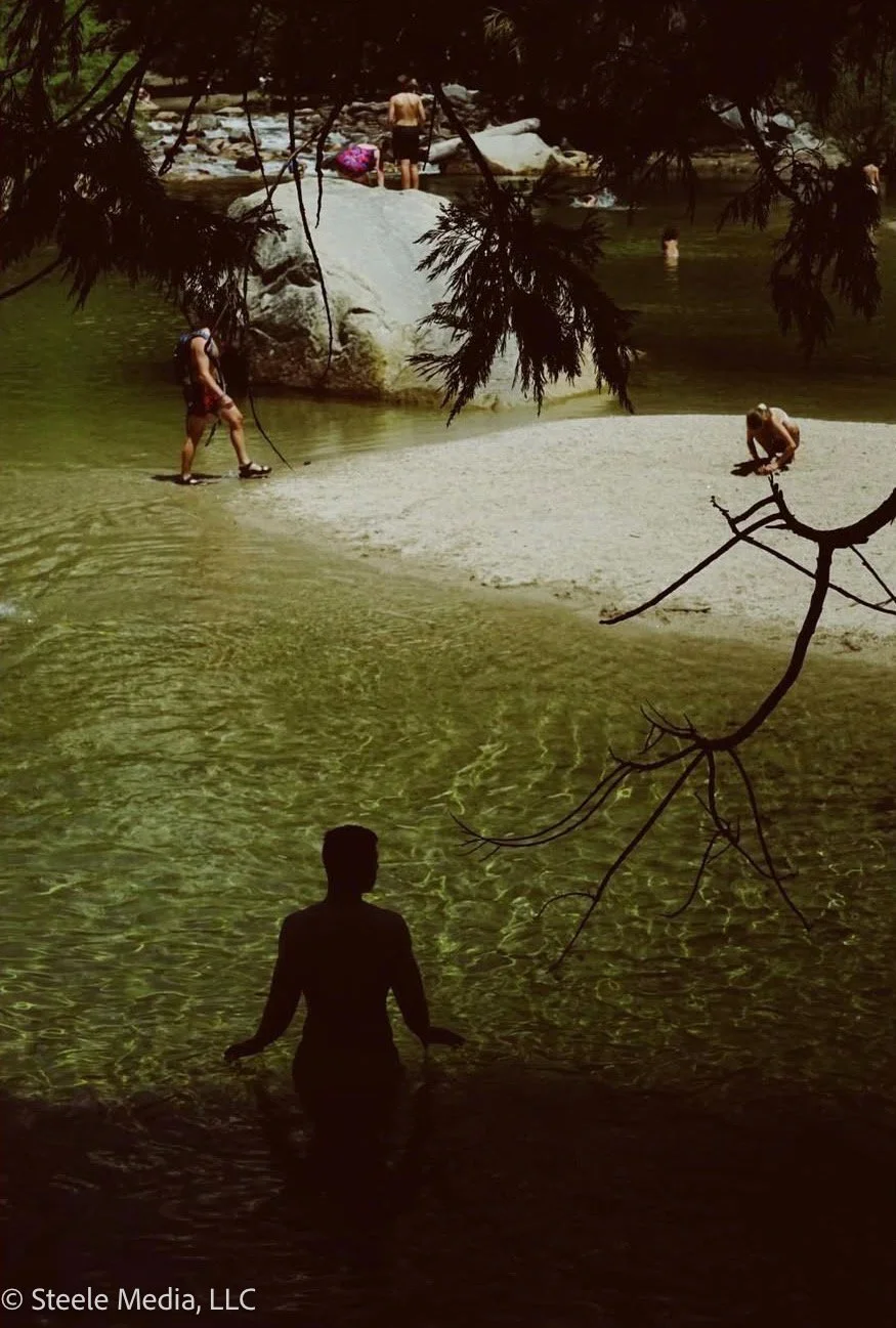 People swimming and relaxing at a river, some sitting on rocks, others wading in the water, surrounded by trees.