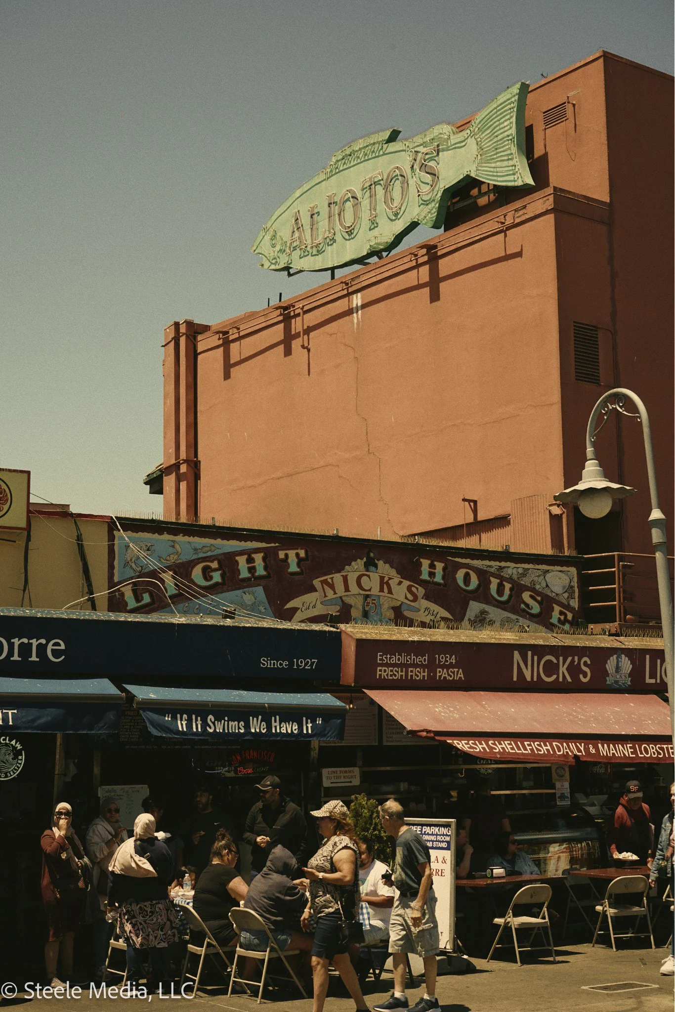 People gathered outside Nick's Fish House restaurant with red awning and signage, next to a pink building with a large fish-shaped sign reading 'Alo Totos'.