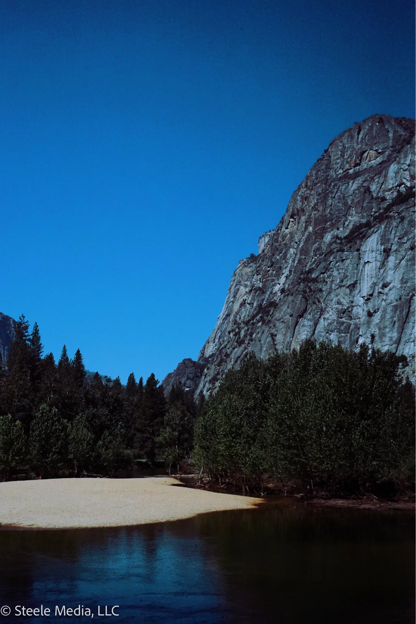A mountain with steep, rocky cliffs next to a dense forest of tall trees, a sandy shore, and a calm river reflecting the landscape under a clear blue sky.