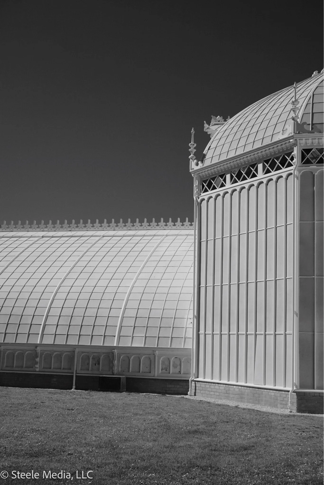 Black and white photo of a Victorian-style glass greenhouse with a curved roof and decorative spires on top, situated outdoors on a grassy area.