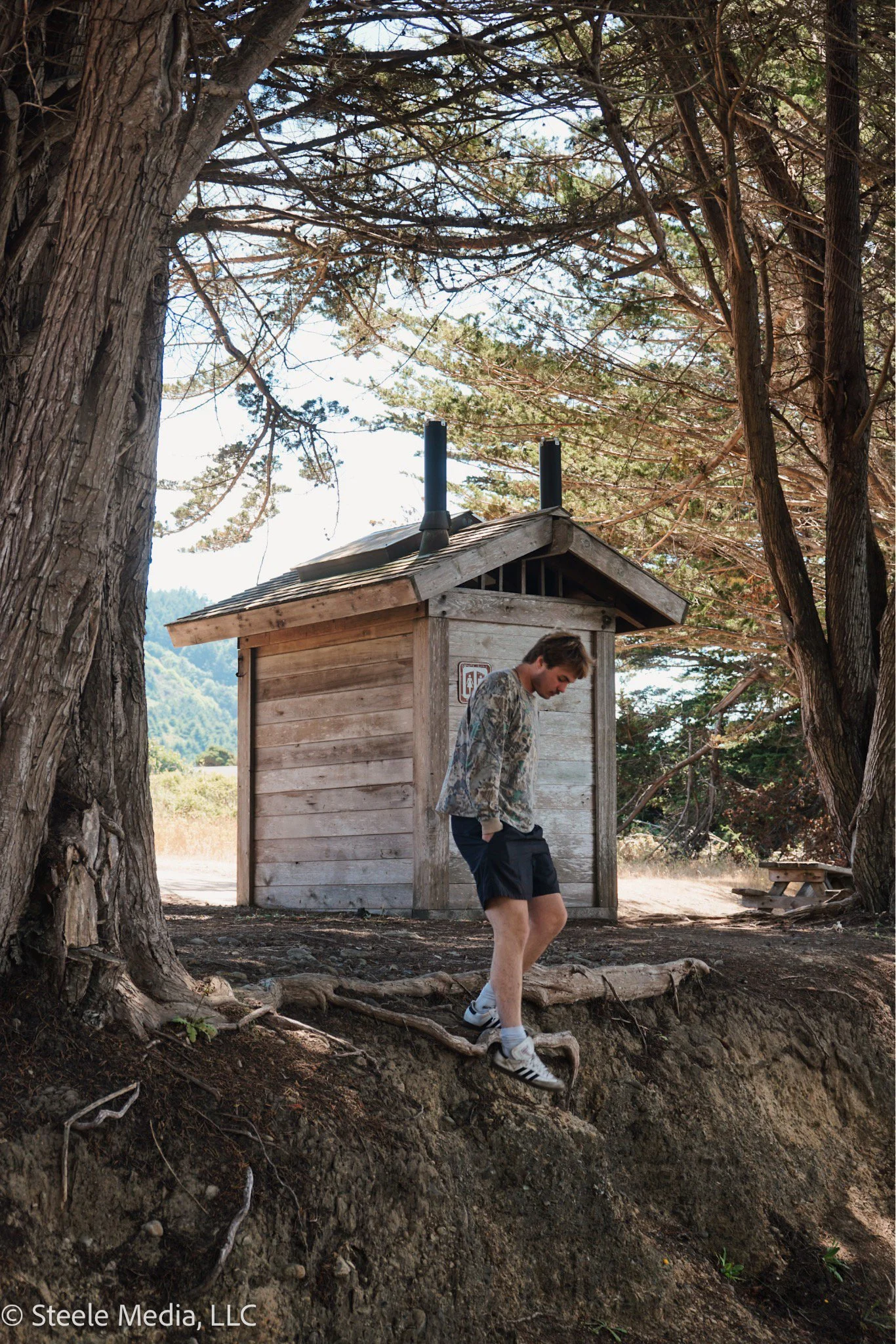 A young man in athletic clothing walks along a dirt path next to a large tree, near a small wooden shed, in a rural outdoor setting with trees and mountains in the background.
