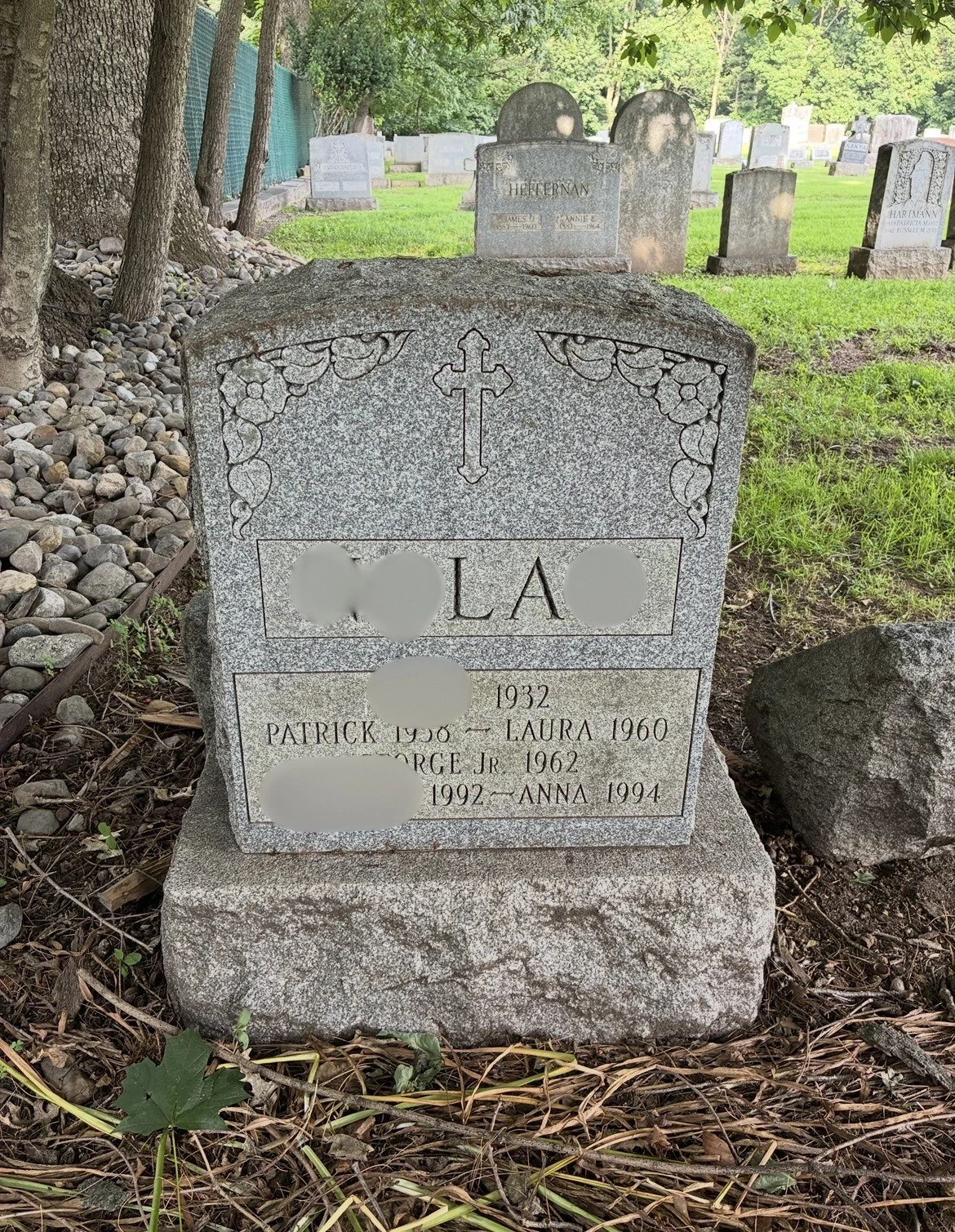 Gravestone in a cemetery with a cross engraved at the top, decorative floral patterns, and inscriptions with names and dates of the deceased. The surrounding area has grass, rocks, and trees.