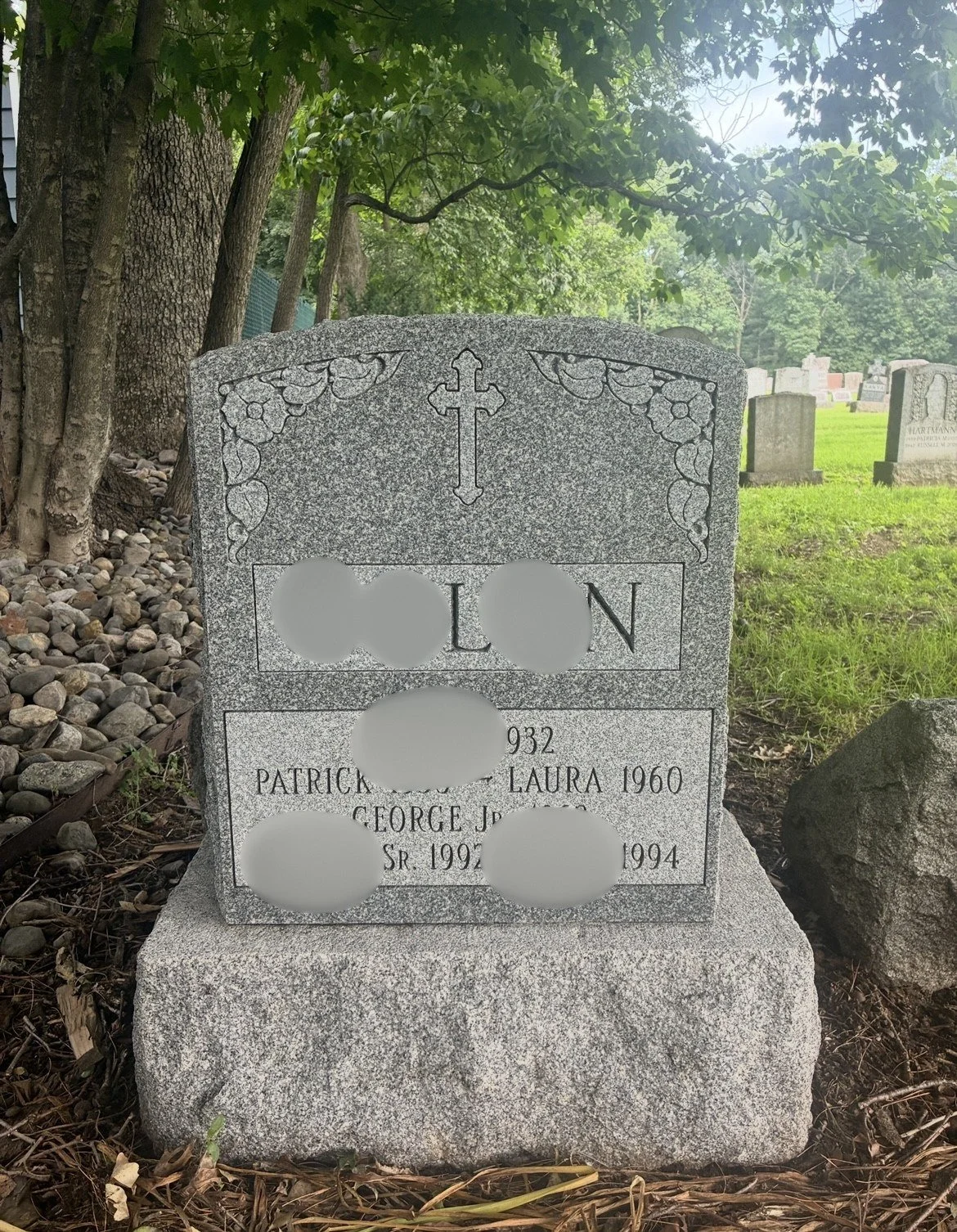 Gravestone in a cemetery with a Christian cross at the top, decorated with carved flowers, and engraved names and dates of deceased individuals. The background shows other gravestones, trees, and green grass.